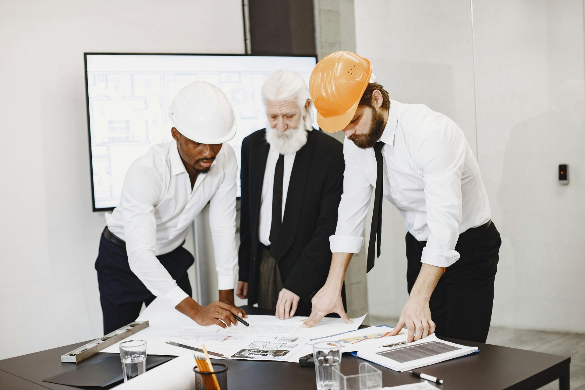 Three workers in hard hats review blueprints at a conference table in an office.