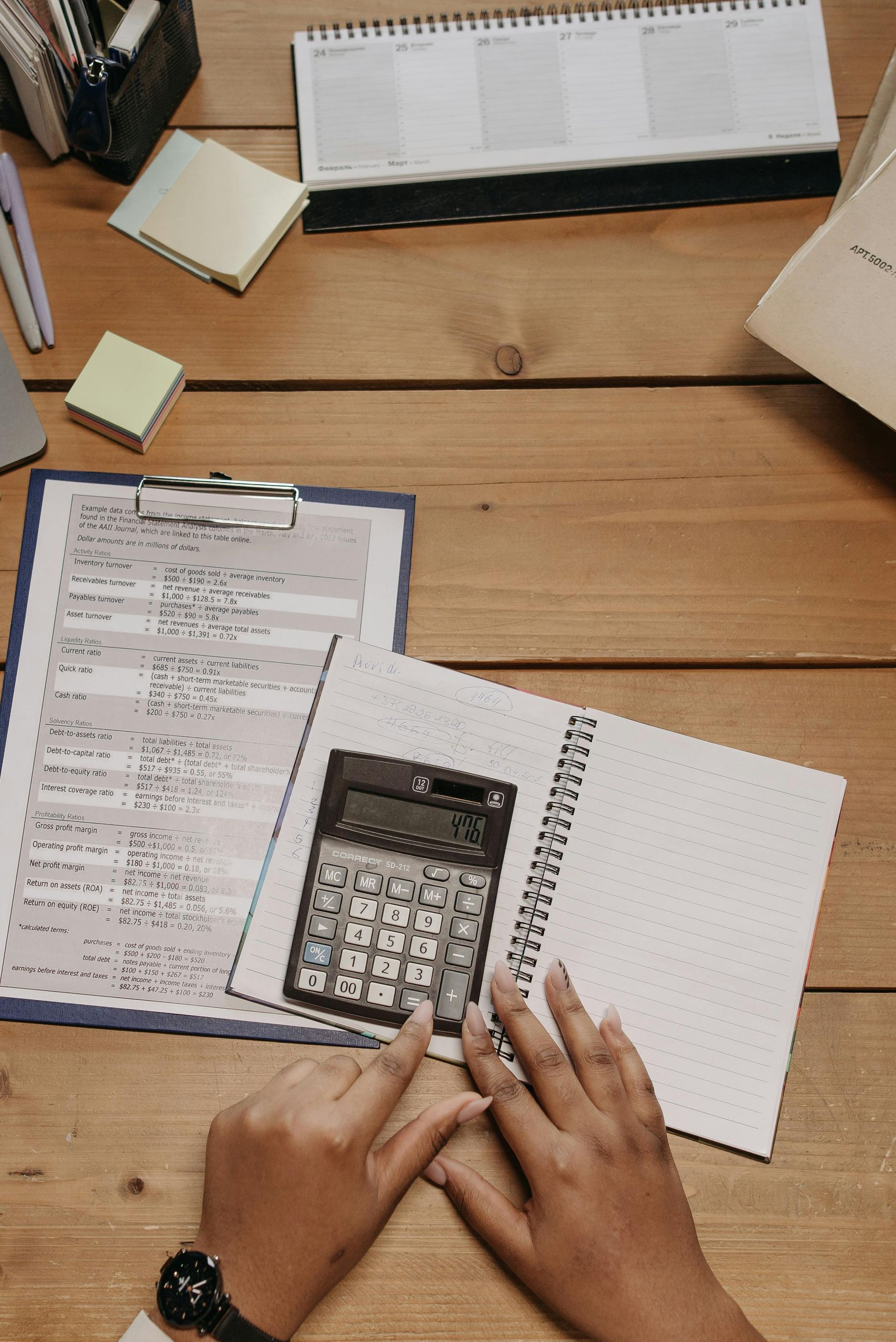 Hands using a calculator and open notebook on a wooden desk with papers and sticky notes