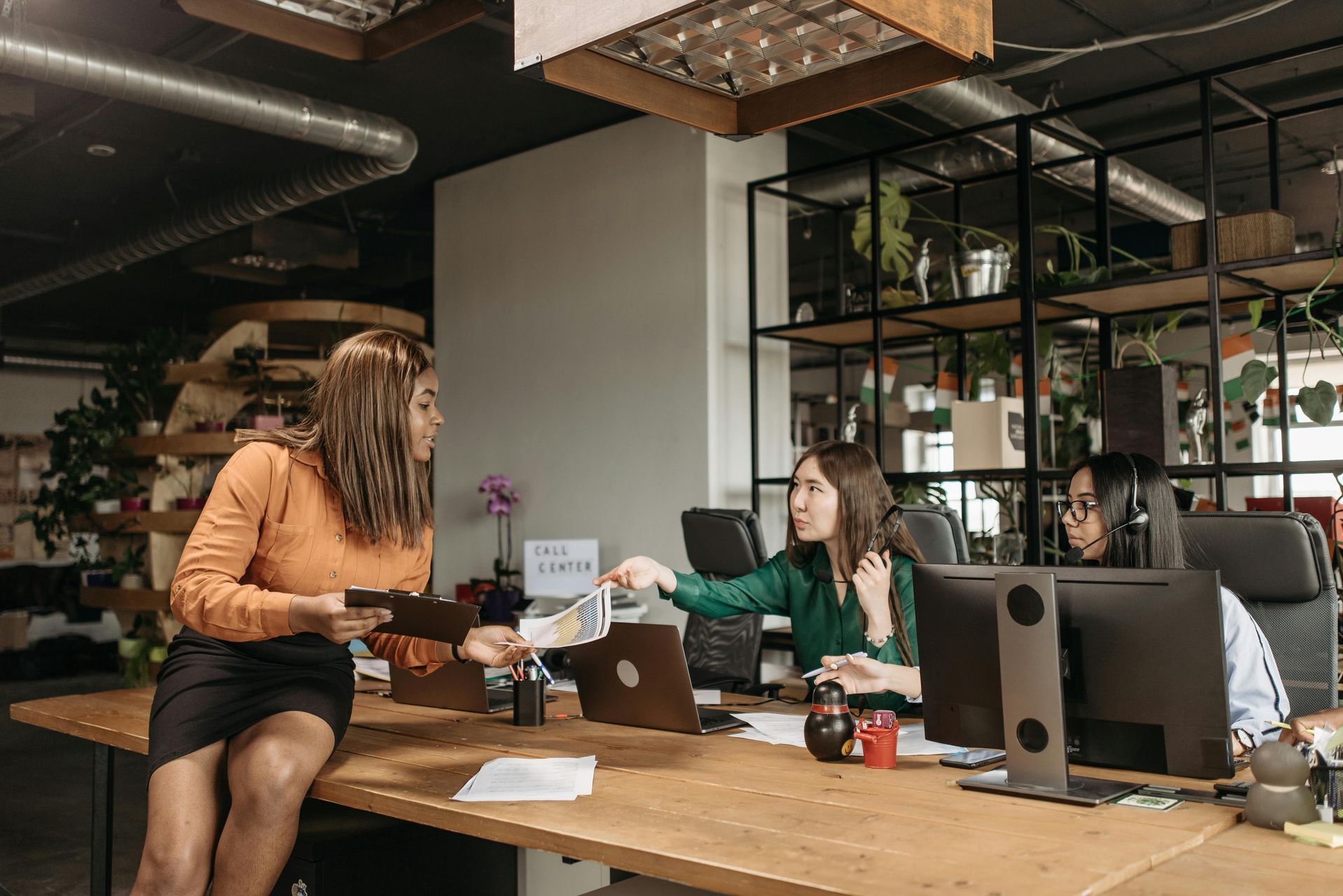 Two coworkers exchanging a card across a desk in a modern office with computers and plants.