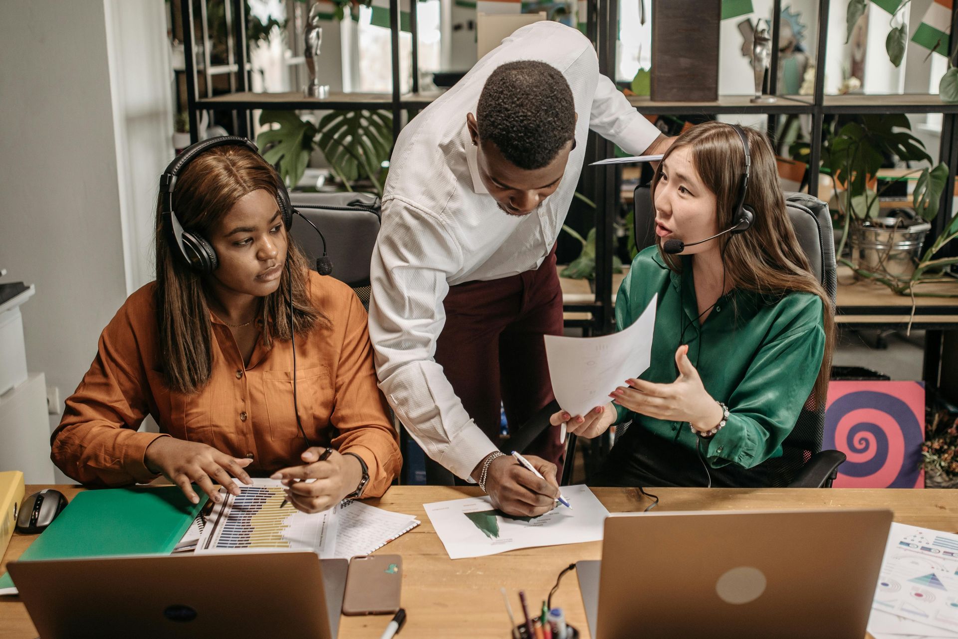 Three colleagues collaborate at a table with laptops and papers in a bright office.