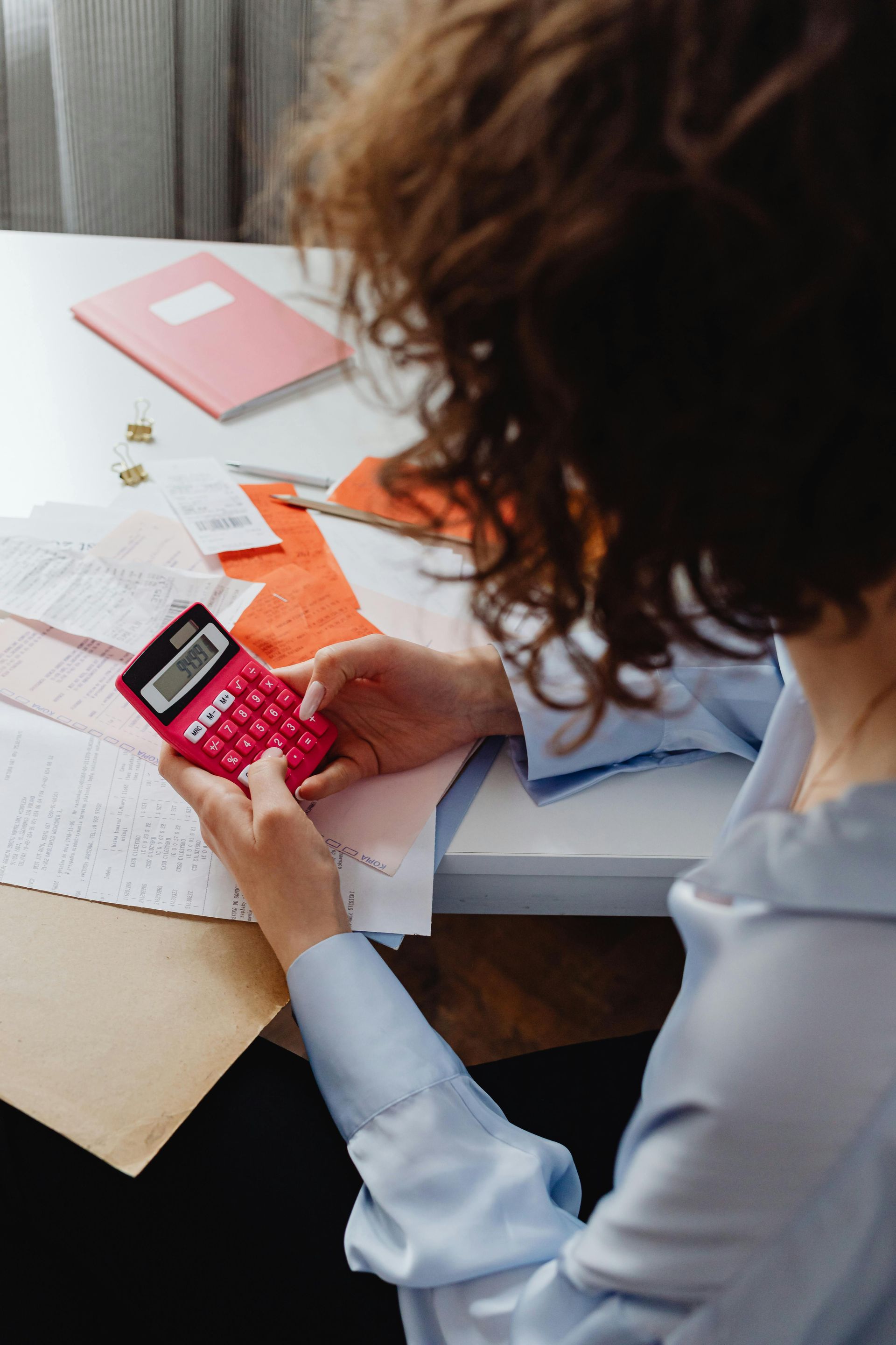 Person using a pink calculator while reviewing papers at a desk with a notebook and laptop