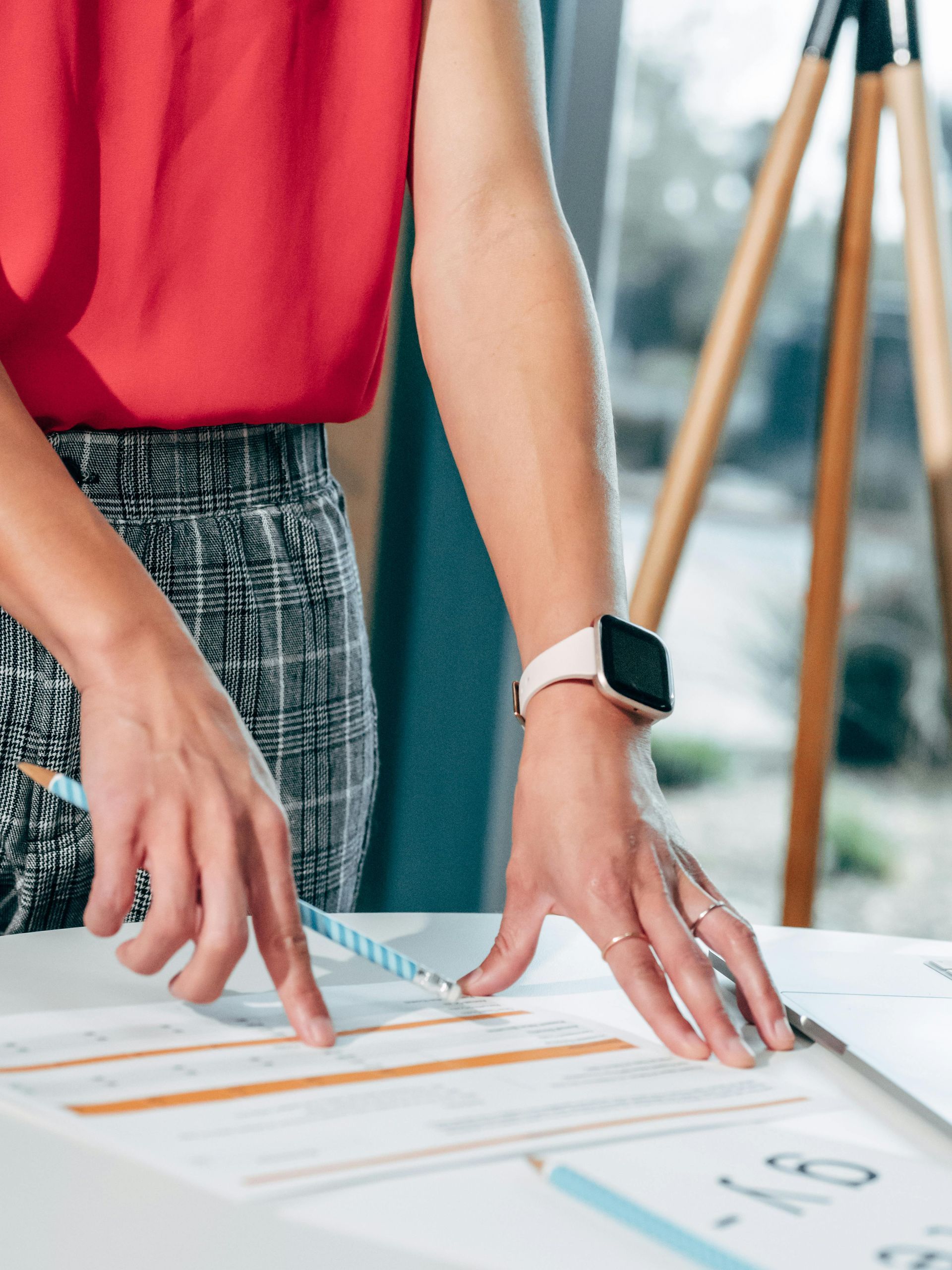 Person pointing at charts on a desk, wearing a red top and smartwatch.