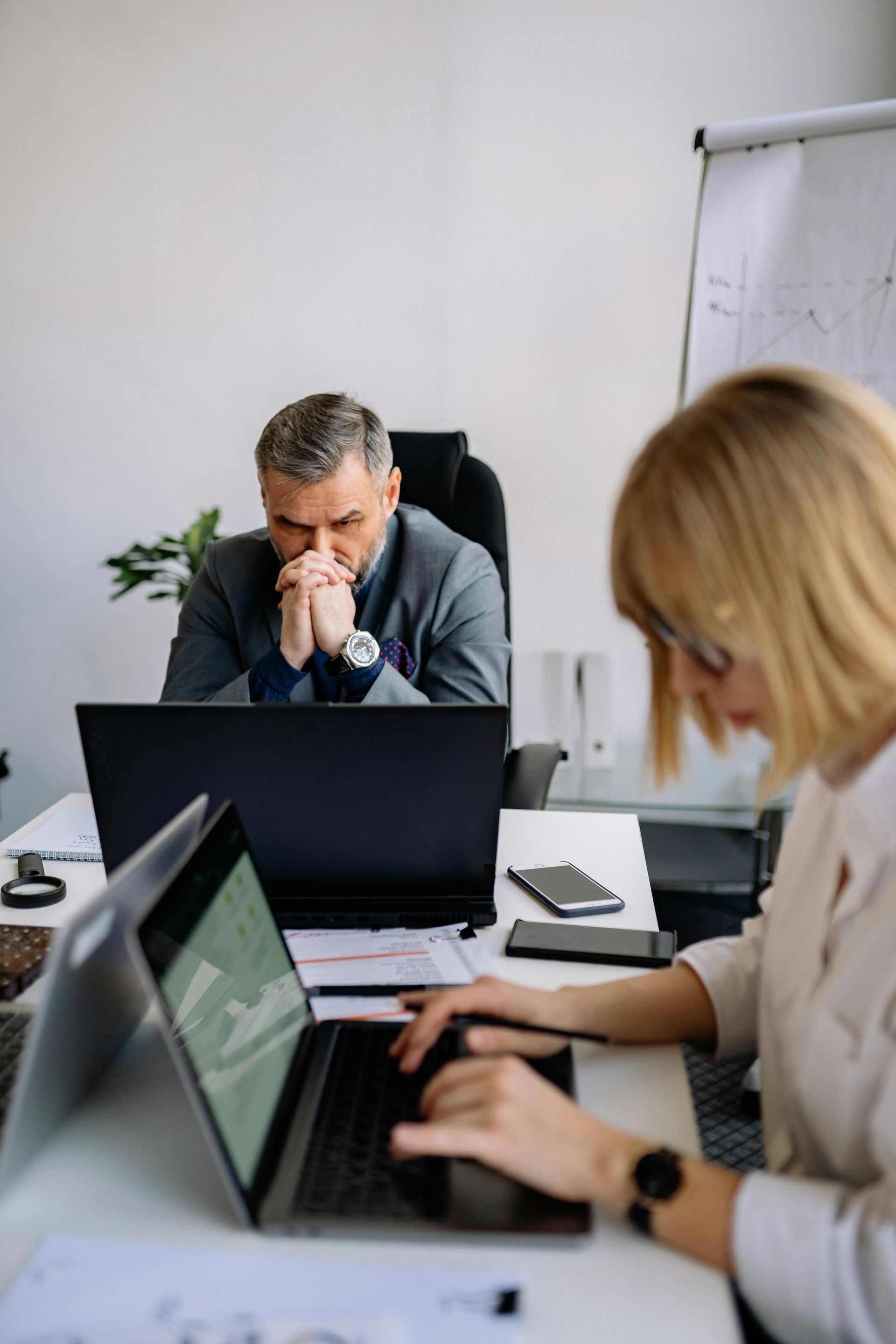 Two people working on laptops in a meeting room, with a man thinking at the table and a whiteboard behind them.