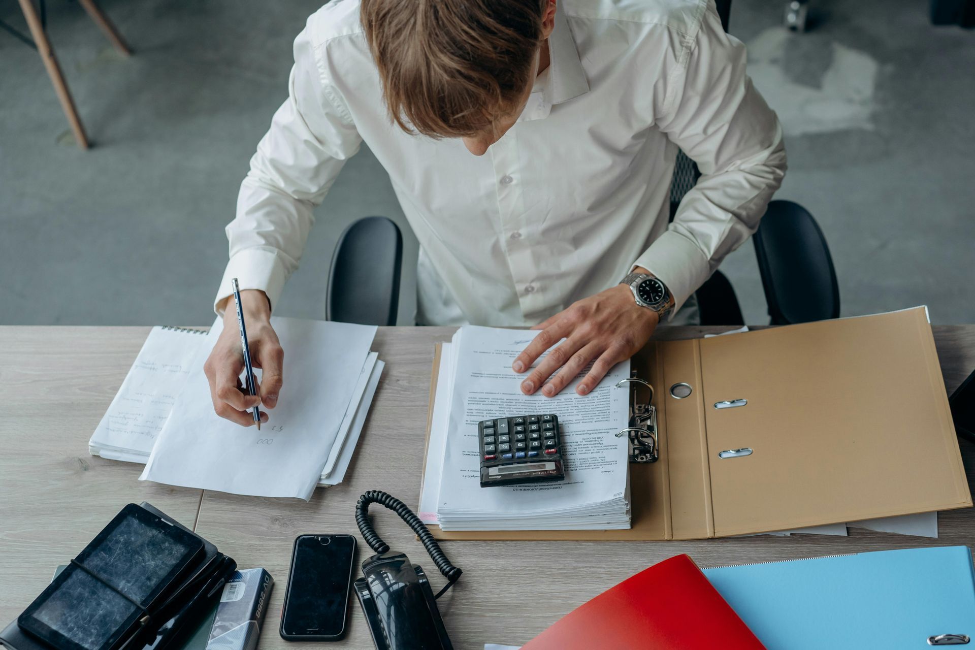 Person working at a desk, writing in a notebook and using a calculator beside files and stationery.