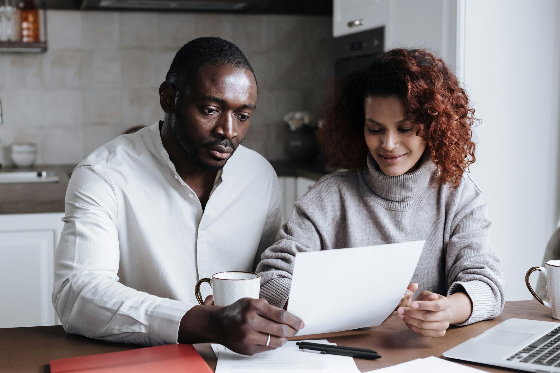 Two people reviewing a document together at a table in a kitchen, with a laptop nearby.