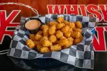 Basket of golden fried tater tots with dipping sauce on black-and-white checkered paper