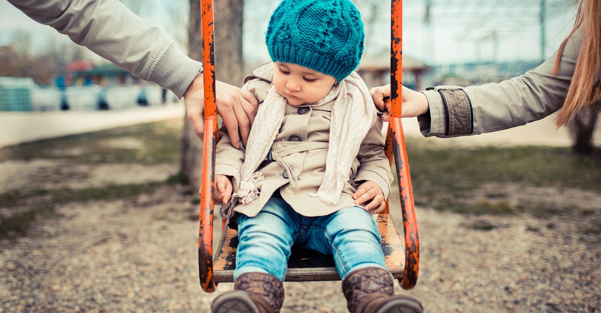 A baby is sitting on a swing being held by two people.