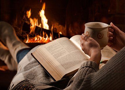 A person is sitting in front of a fireplace reading a book and drinking coffee.