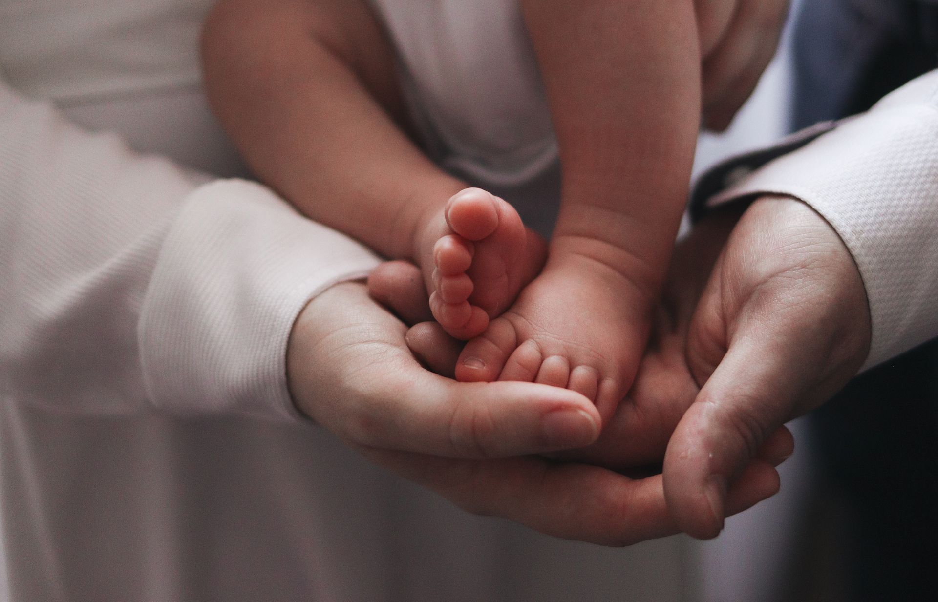 Two adults gently holding a baby’s feet and hands in a soft, close-up scene