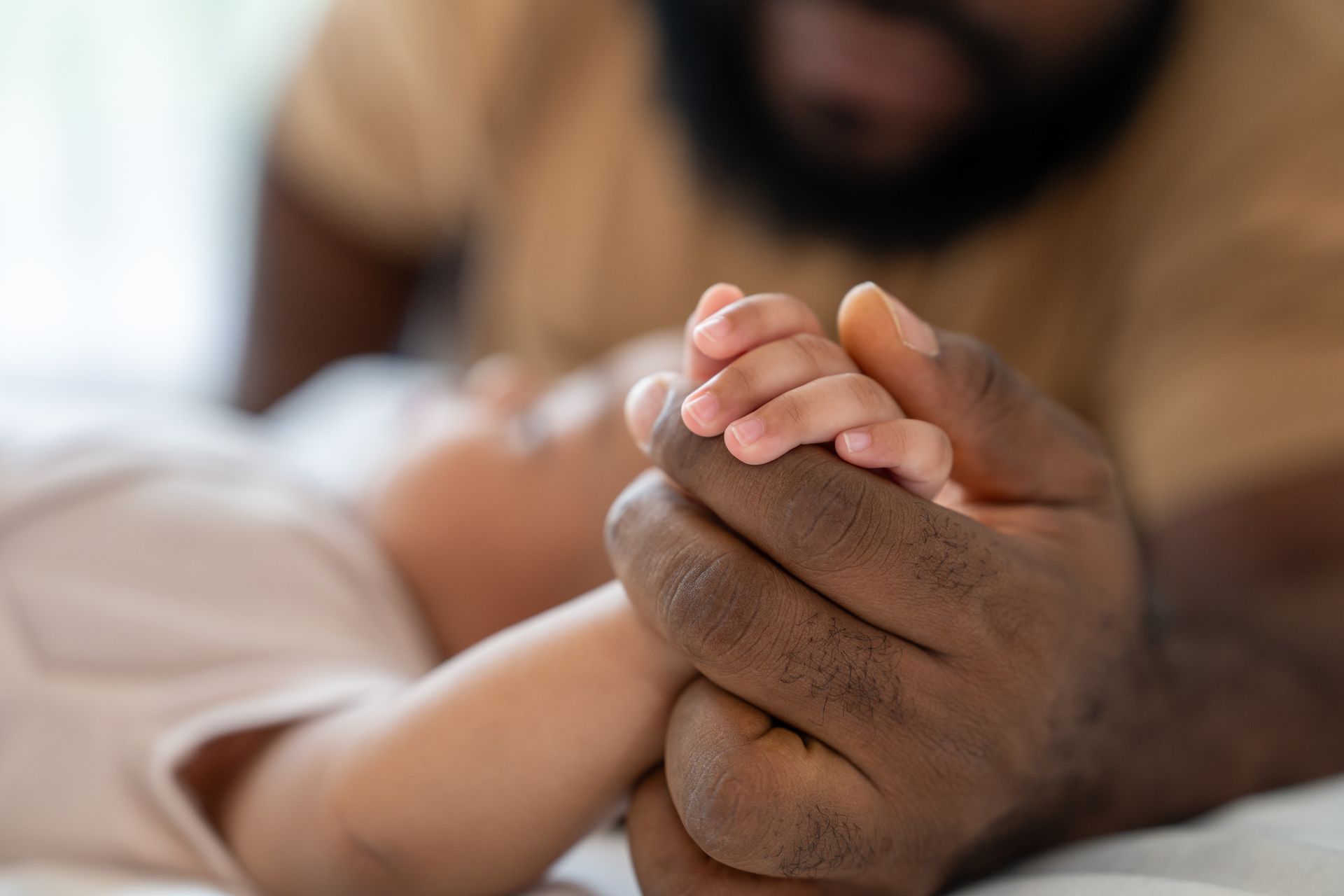 Hands clasped in a close-up on a bed, with a blurred person leaning in the background.