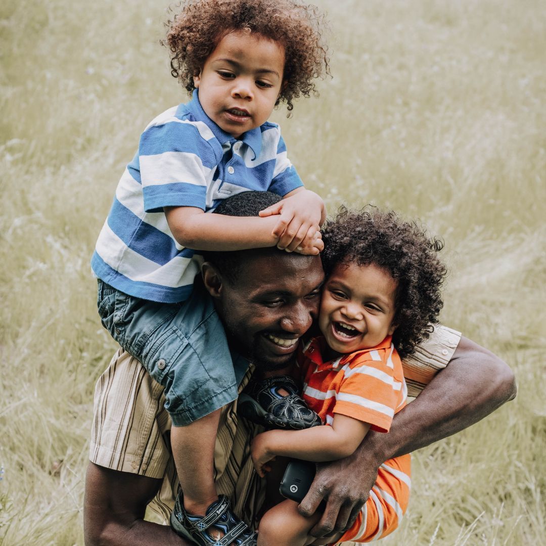 A man is carrying two children on his shoulders in the grass.