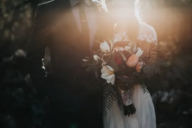 A couple in formal wedding attire stands in soft sunlight, holding a bouquet of white flowers and ferns.