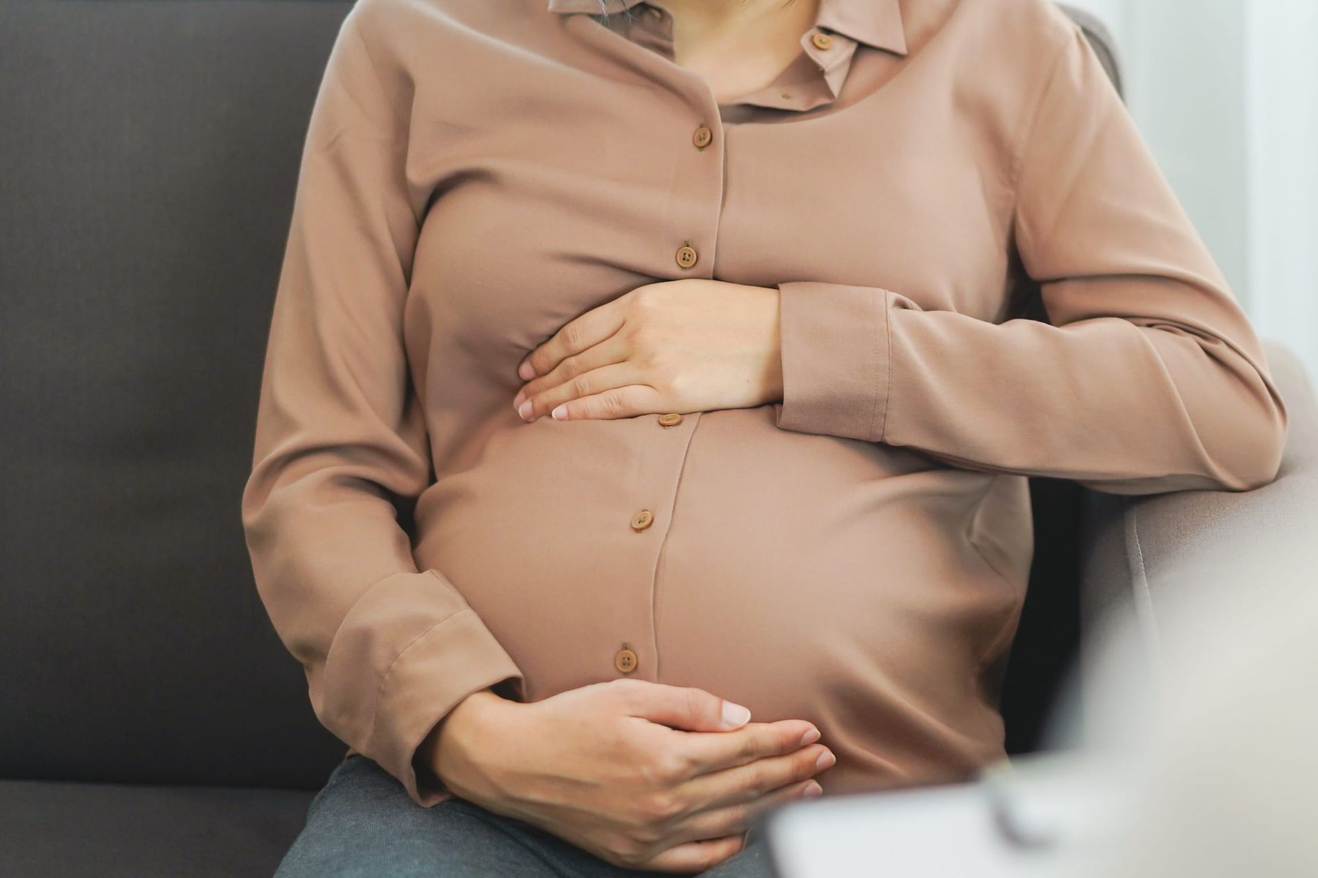 Pregnant person resting hands on belly while seated on a couch, wearing a beige blouse