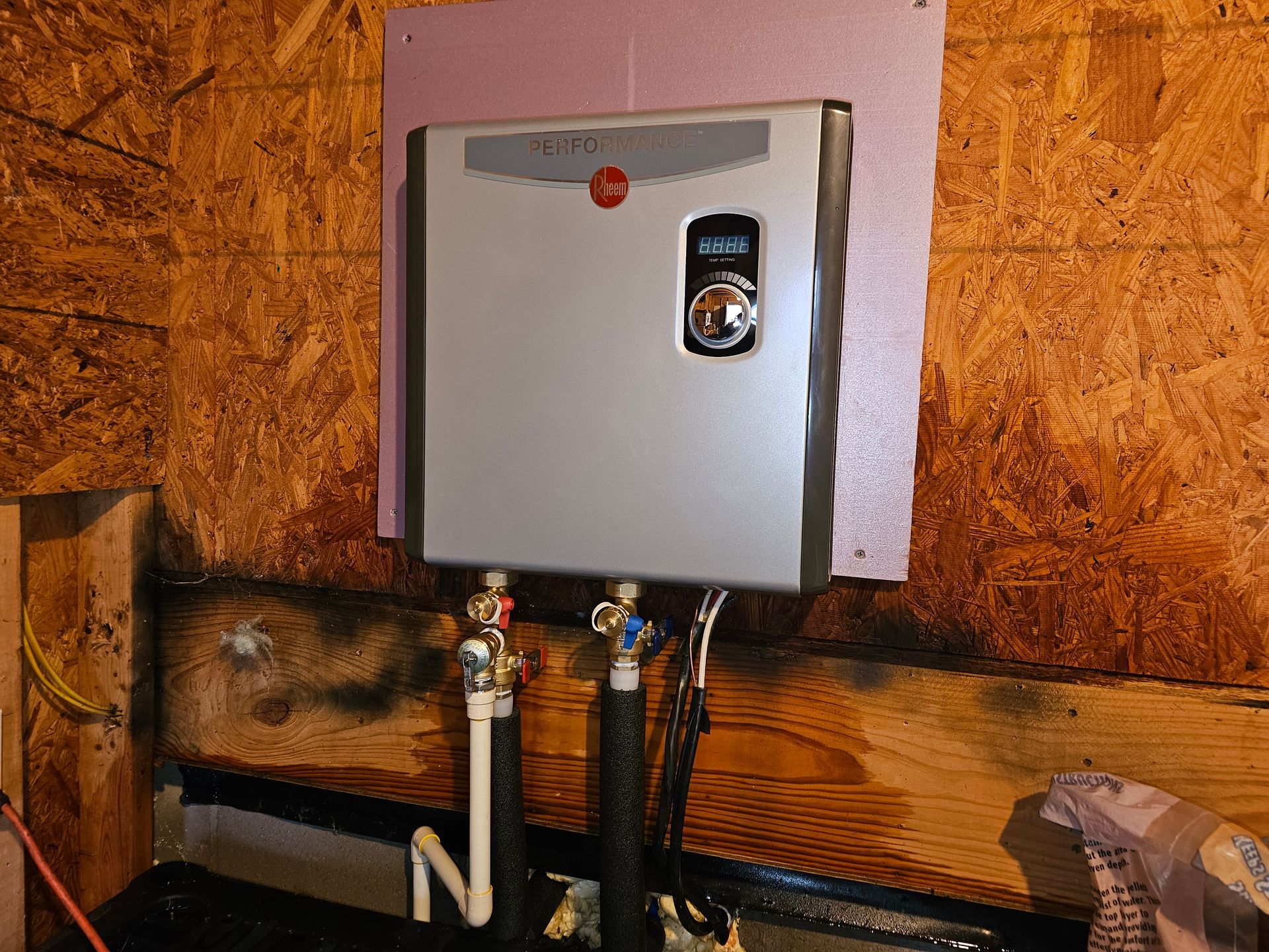 Rheem tankless water heater mounted on wooden wall with pink insulation. Water pipes and controls visible.