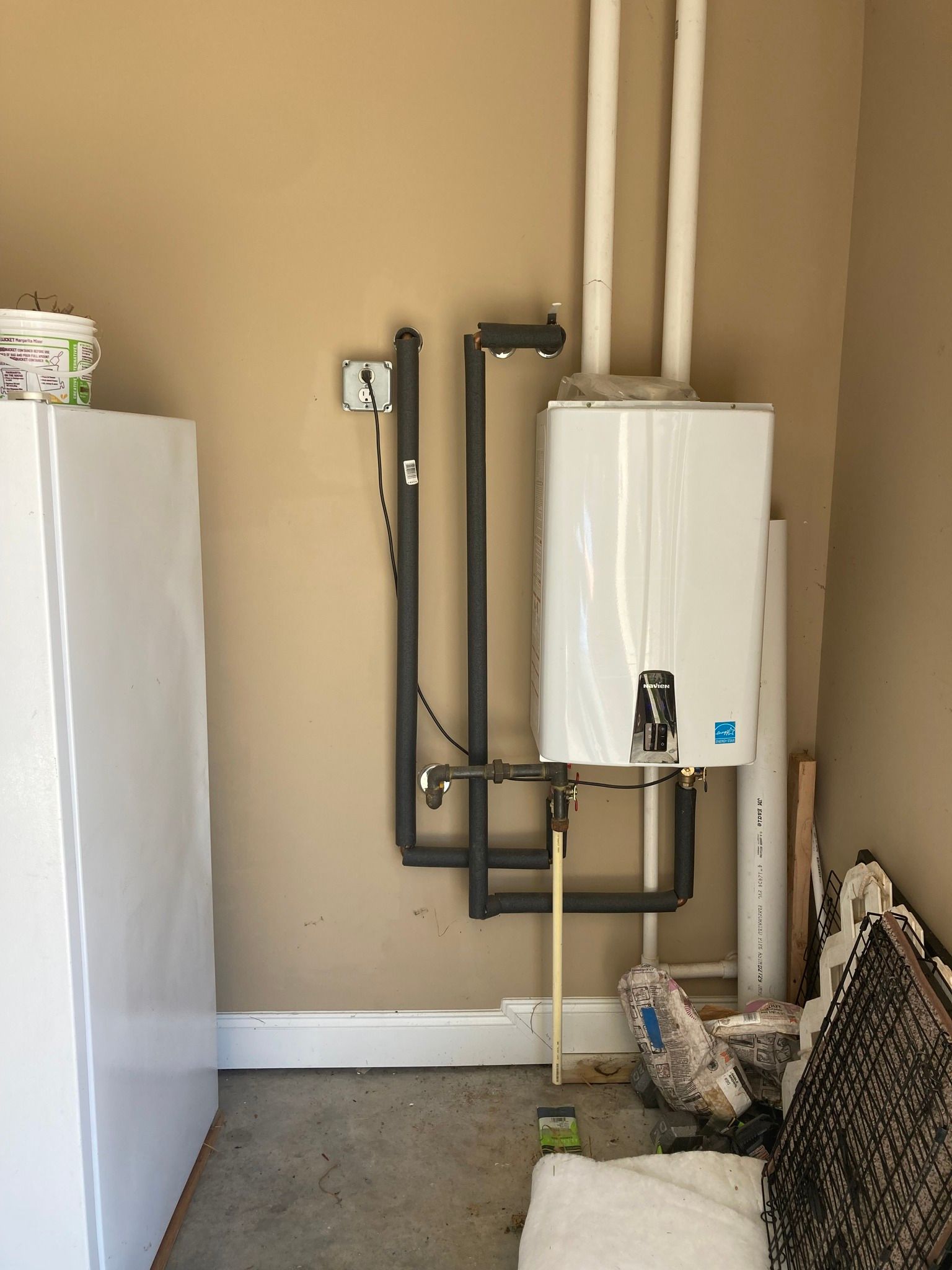 A gas water heater mounted on a tan wall in a garage, with pipes and ventilation.