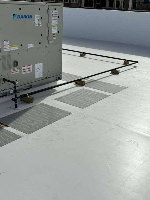 HVAC unit on a white rooftop, supported by wood blocks. Black piping runs along the roof.
