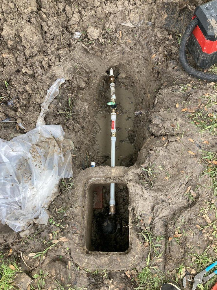 Open ground reveals water pipes and valves, including a meter, in a concrete box.
