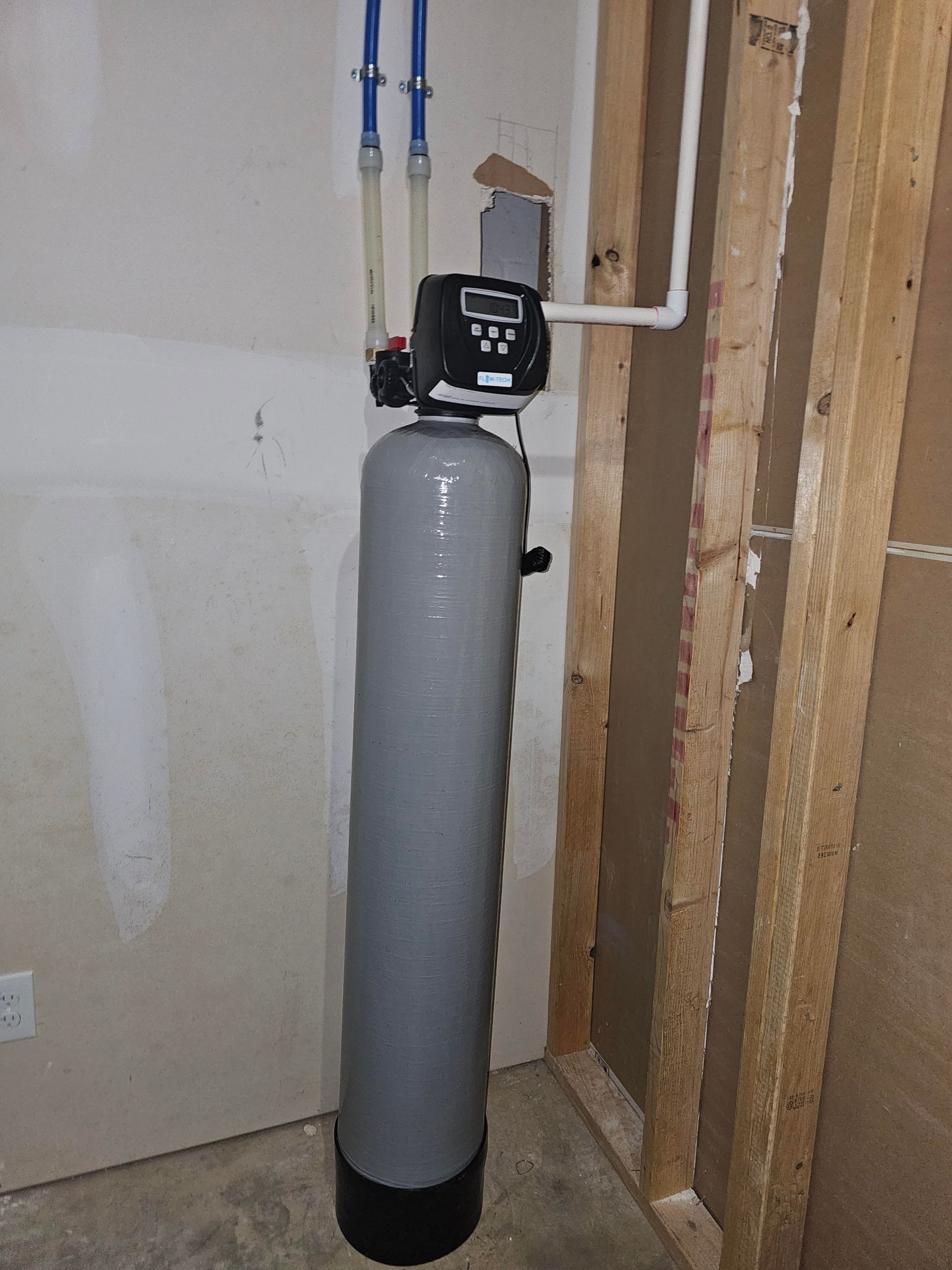 Water softener system in a residential garage, gray tank with digital controls, blue and white pipes.