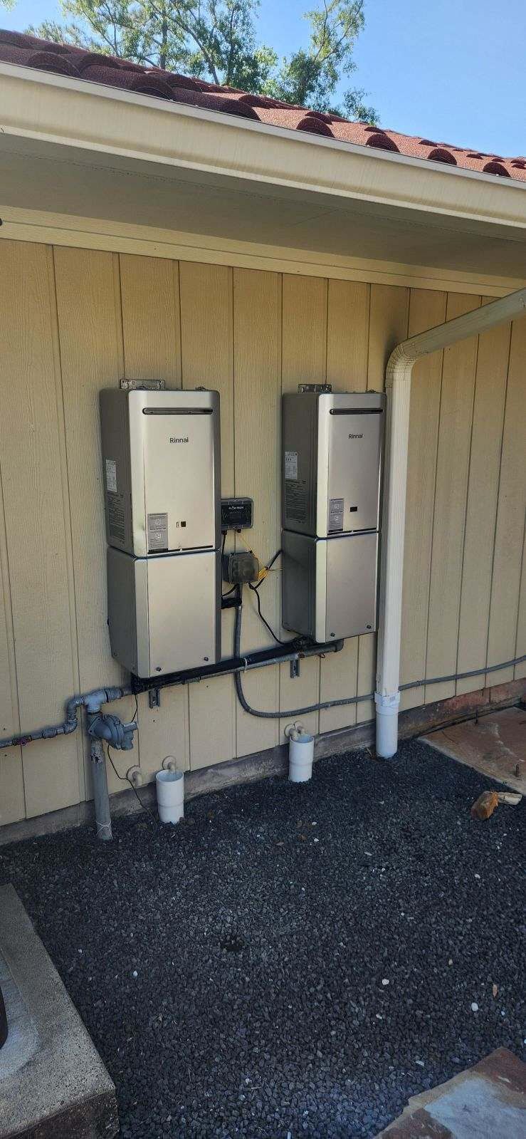 Two silver water heaters mounted on a wall. Piping and gravel ground.