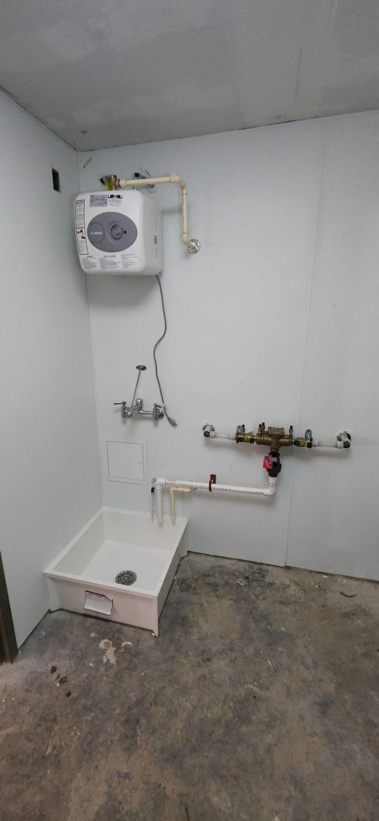 A utility room with a sink, pipes, and a wall-mounted water heater. Concrete floor, white walls.