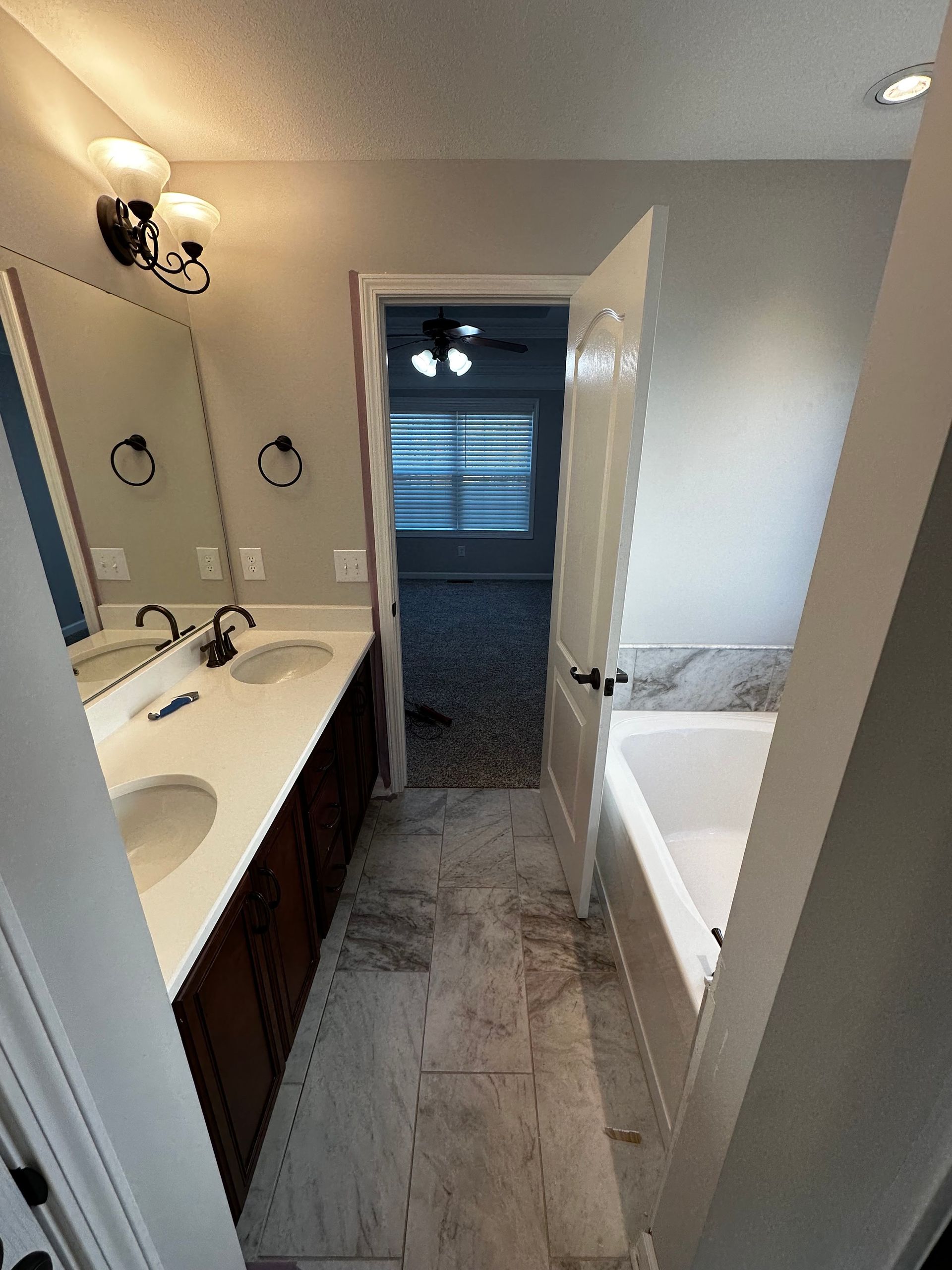 Bathroom with double sinks, tub, and doorway leading to a bedroom. Gray tile floor, light walls.