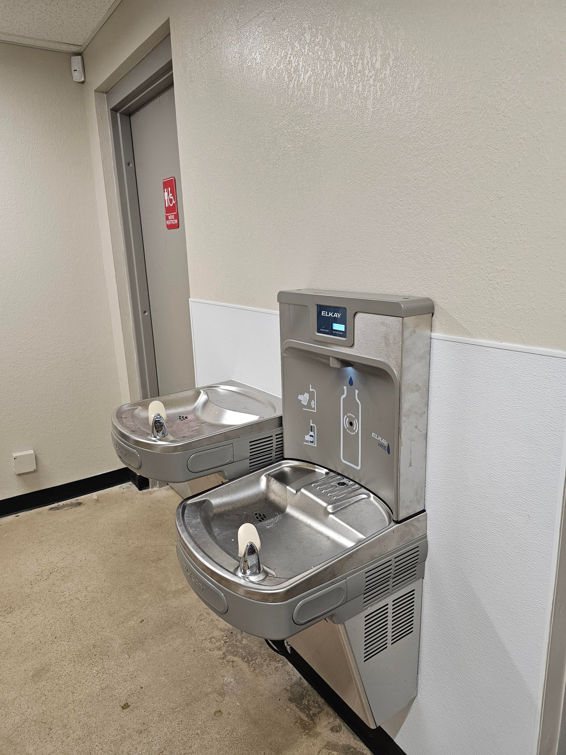 Two stainless steel water fountains mounted on a light-colored wall, with a door in the background.