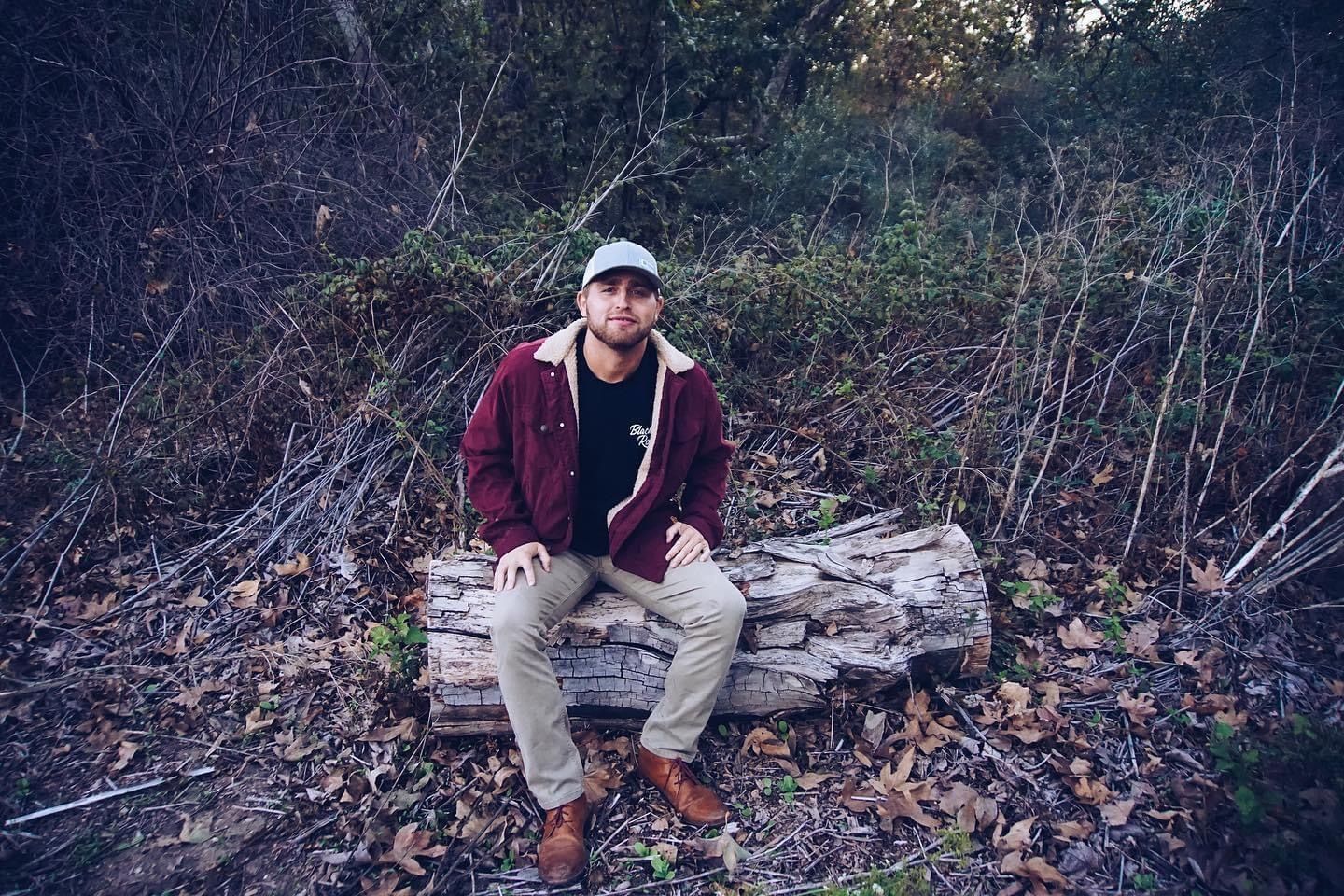 Man in red jacket, cap, sits on a log outdoors.