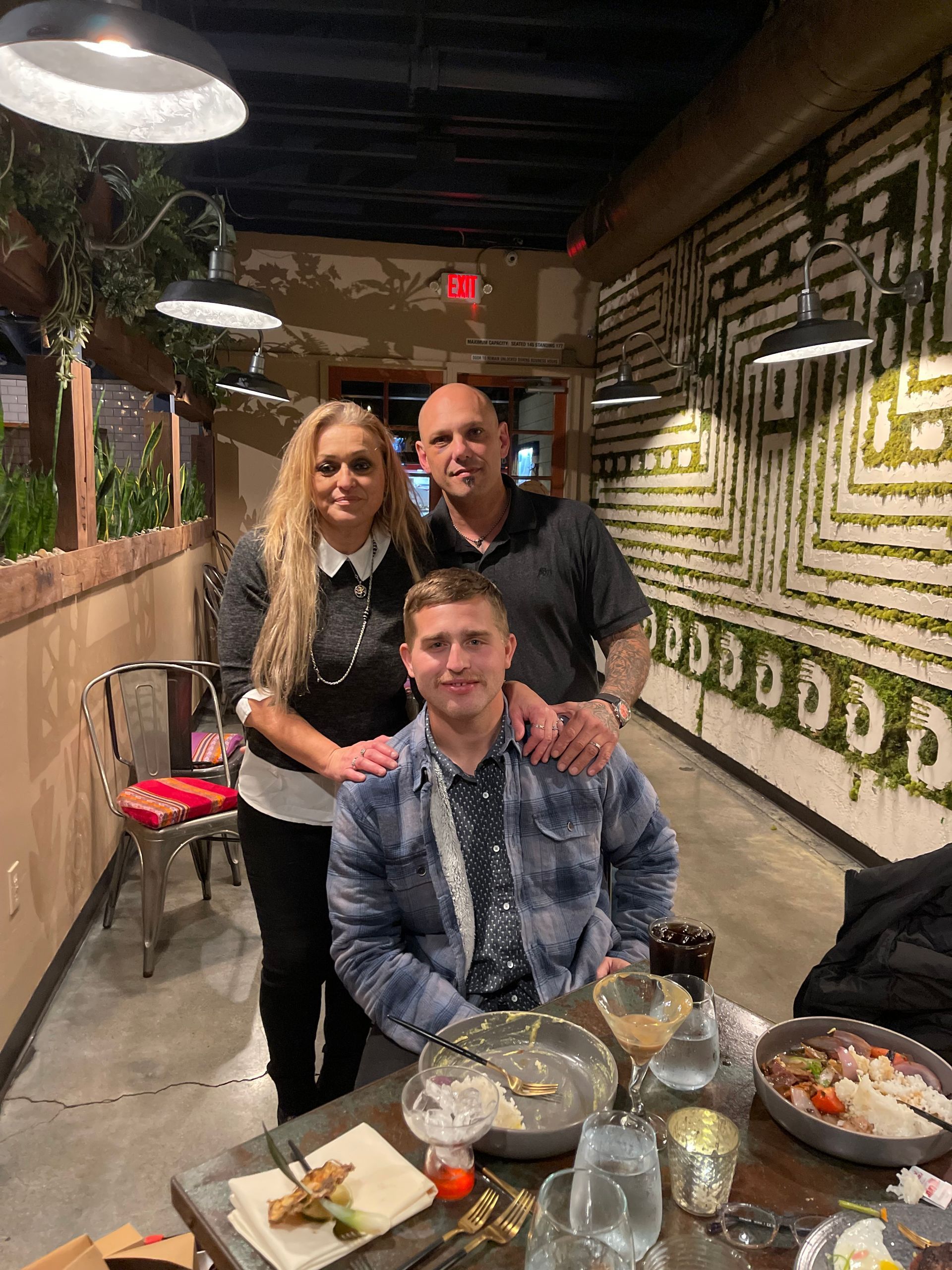 Family of three smiling together at a restaurant, near a decorated wall and table.