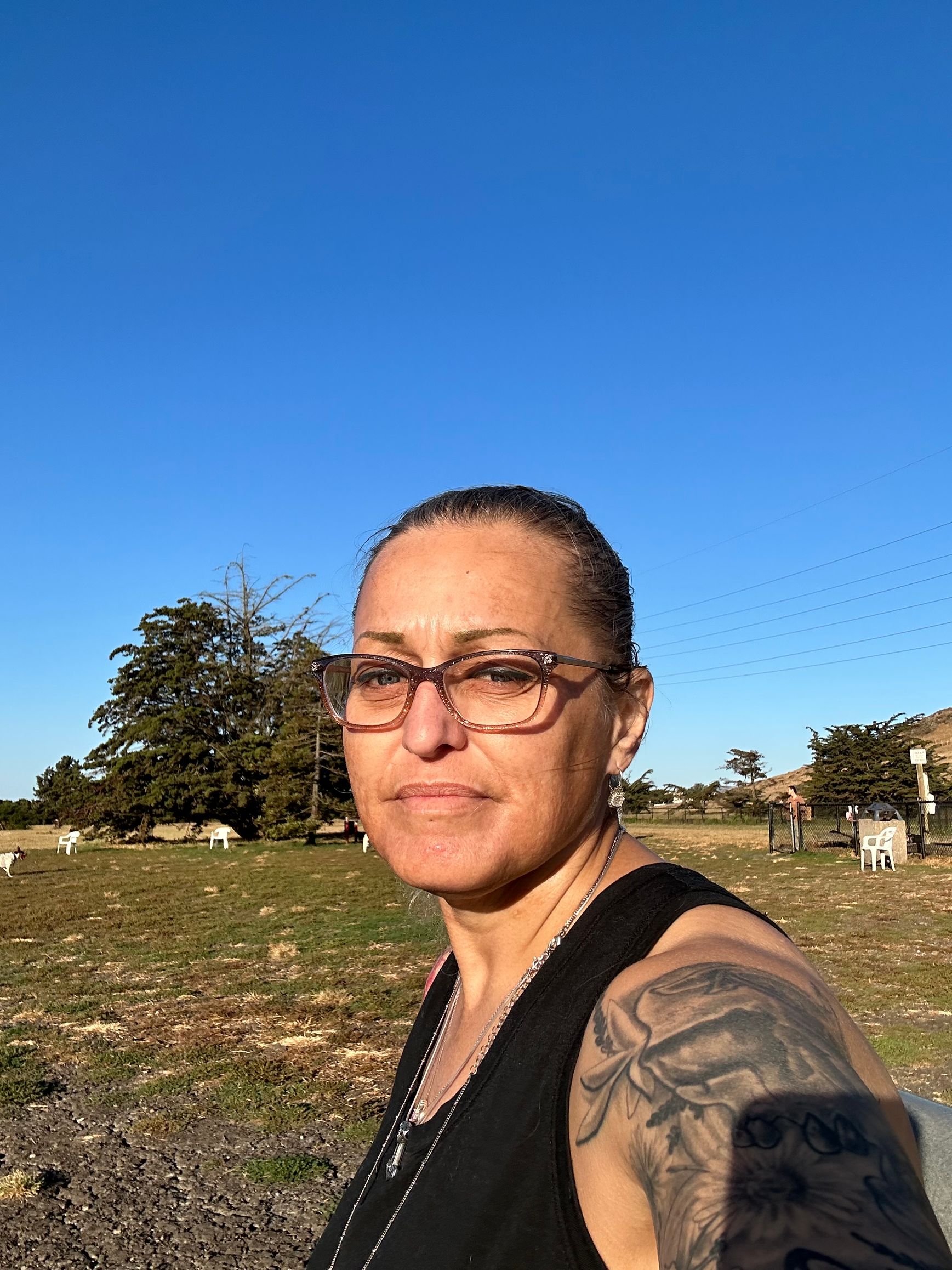 A woman wearing glasses and a tattoo is taking a selfie in a field.