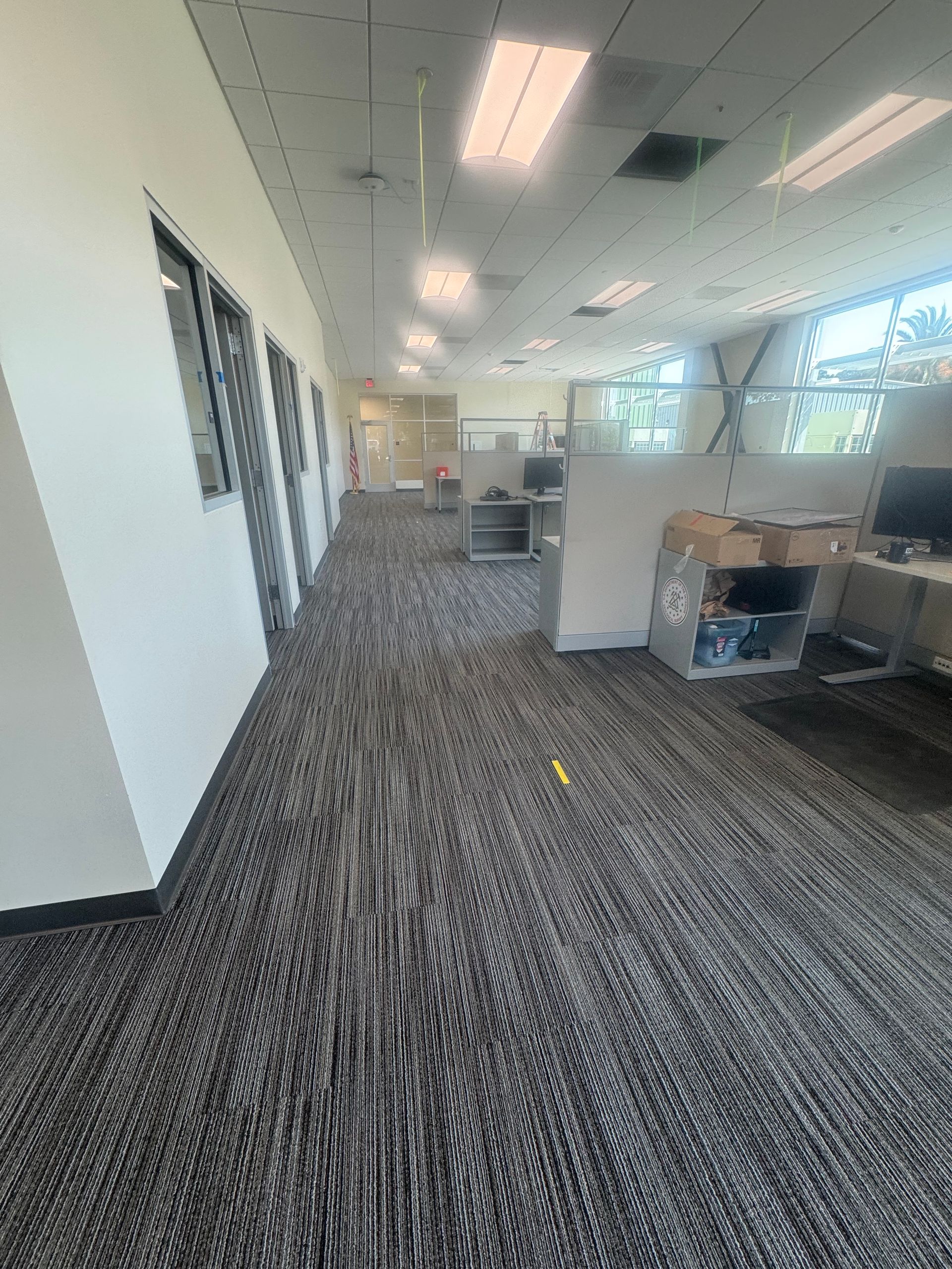 Office hallway with gray carpet, cubicles, and windows.