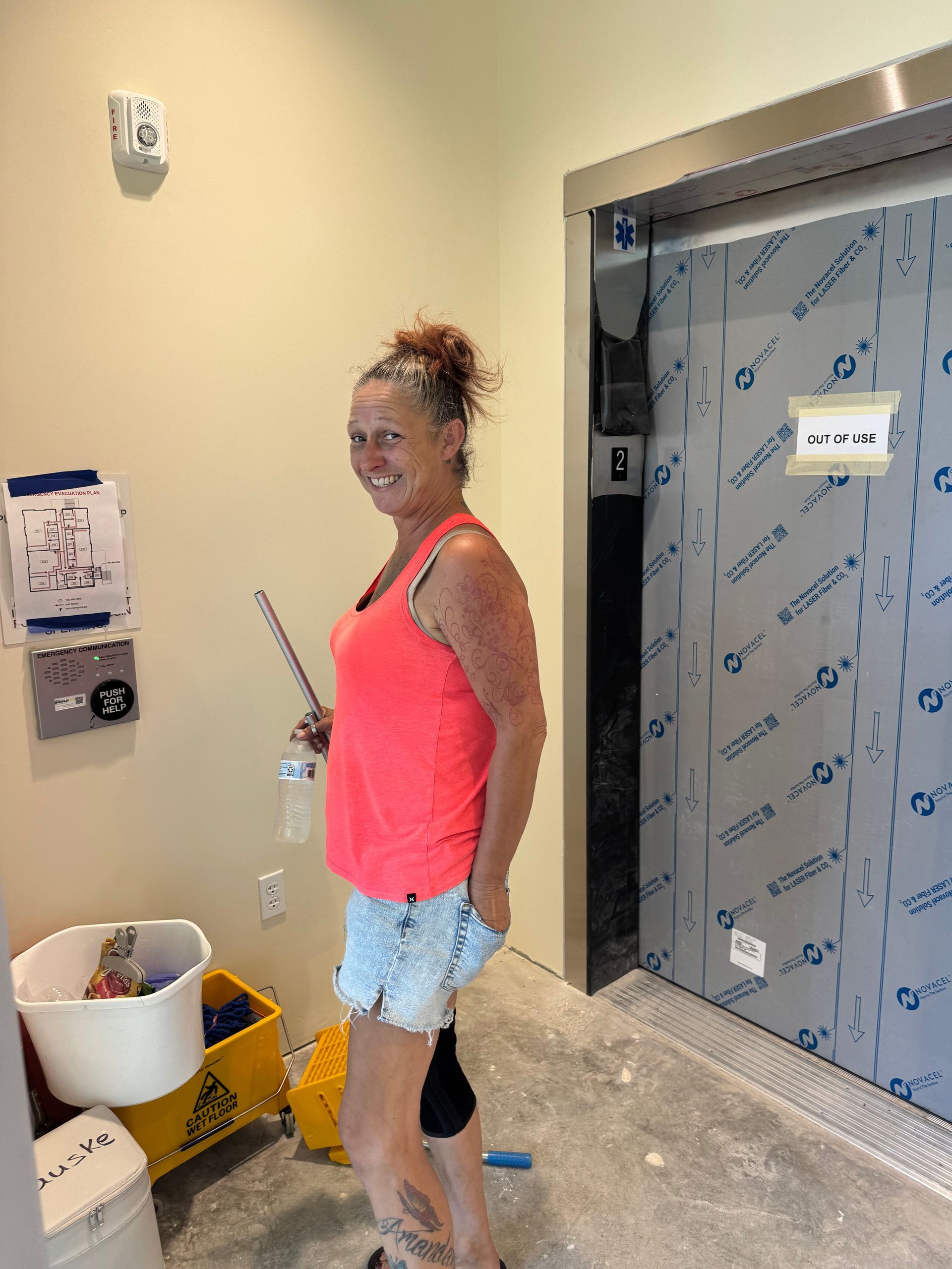 Woman in orange top and shorts smiles near a construction elevator door.