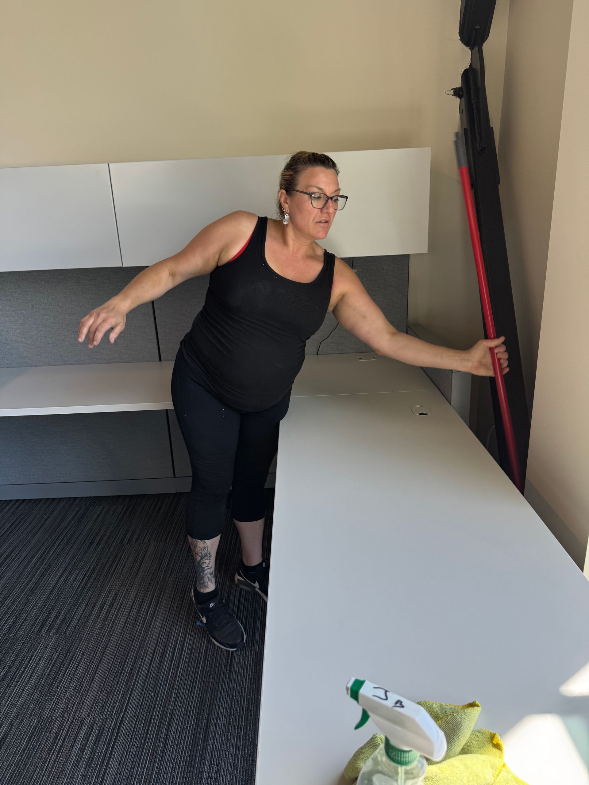 Woman in black activewear, reaching toward a tall, folded object. Room has white desks, cleaning supplies.