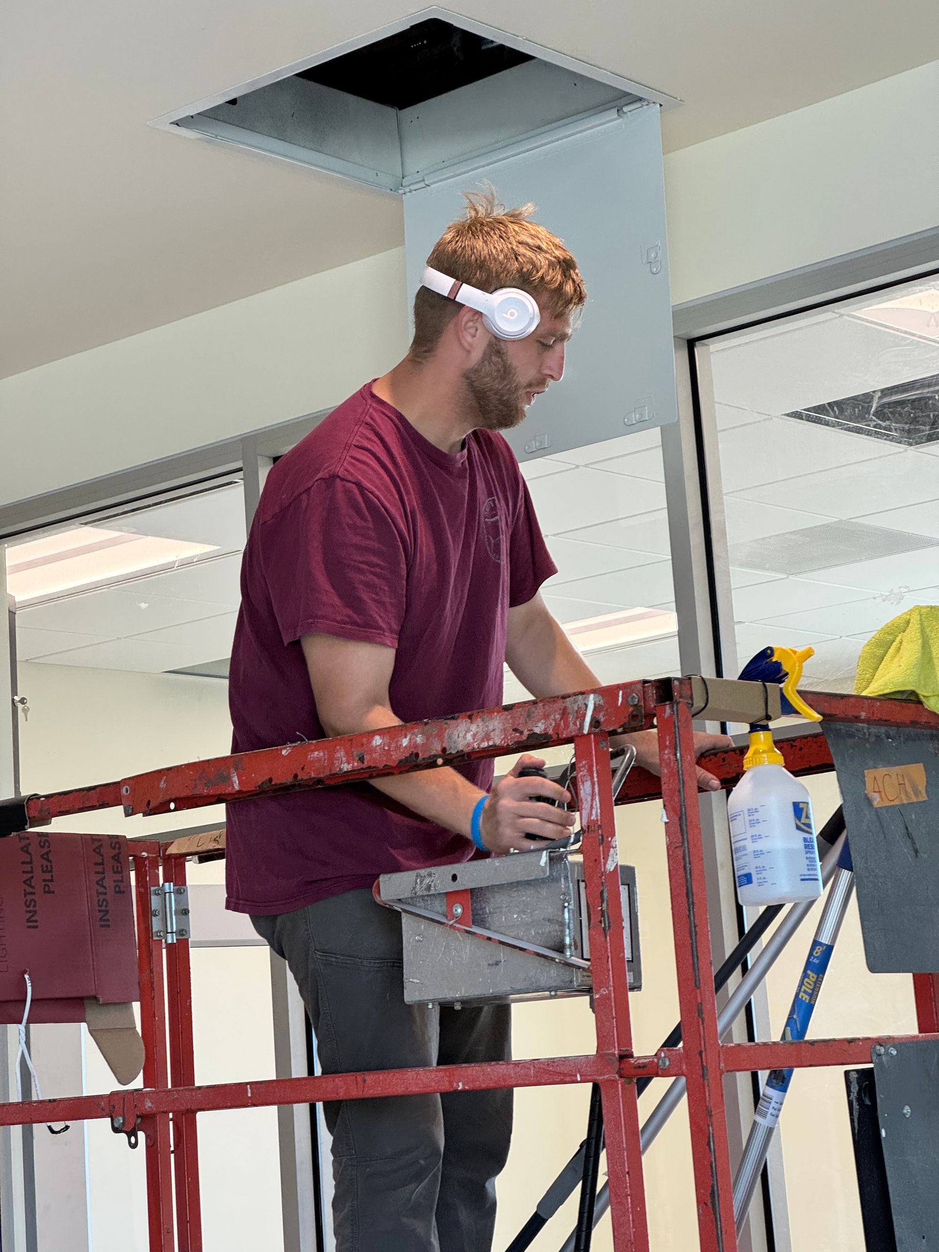Man on a lift cutting into a ceiling. He wears headphones and a maroon shirt, working in a building.