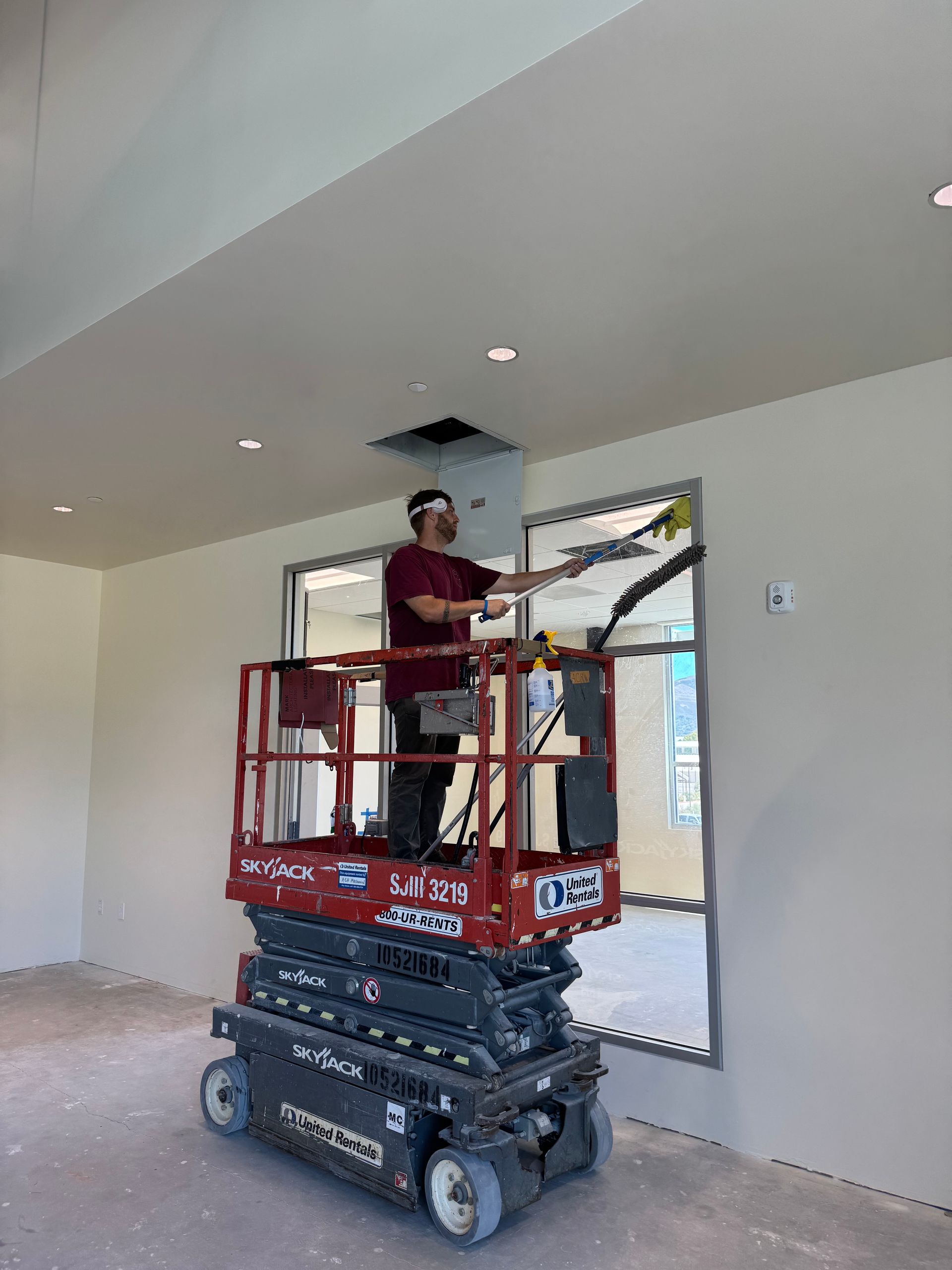 Man on a scissor lift cleans a ceiling vent in a room with neutral-colored walls, using a long tool.