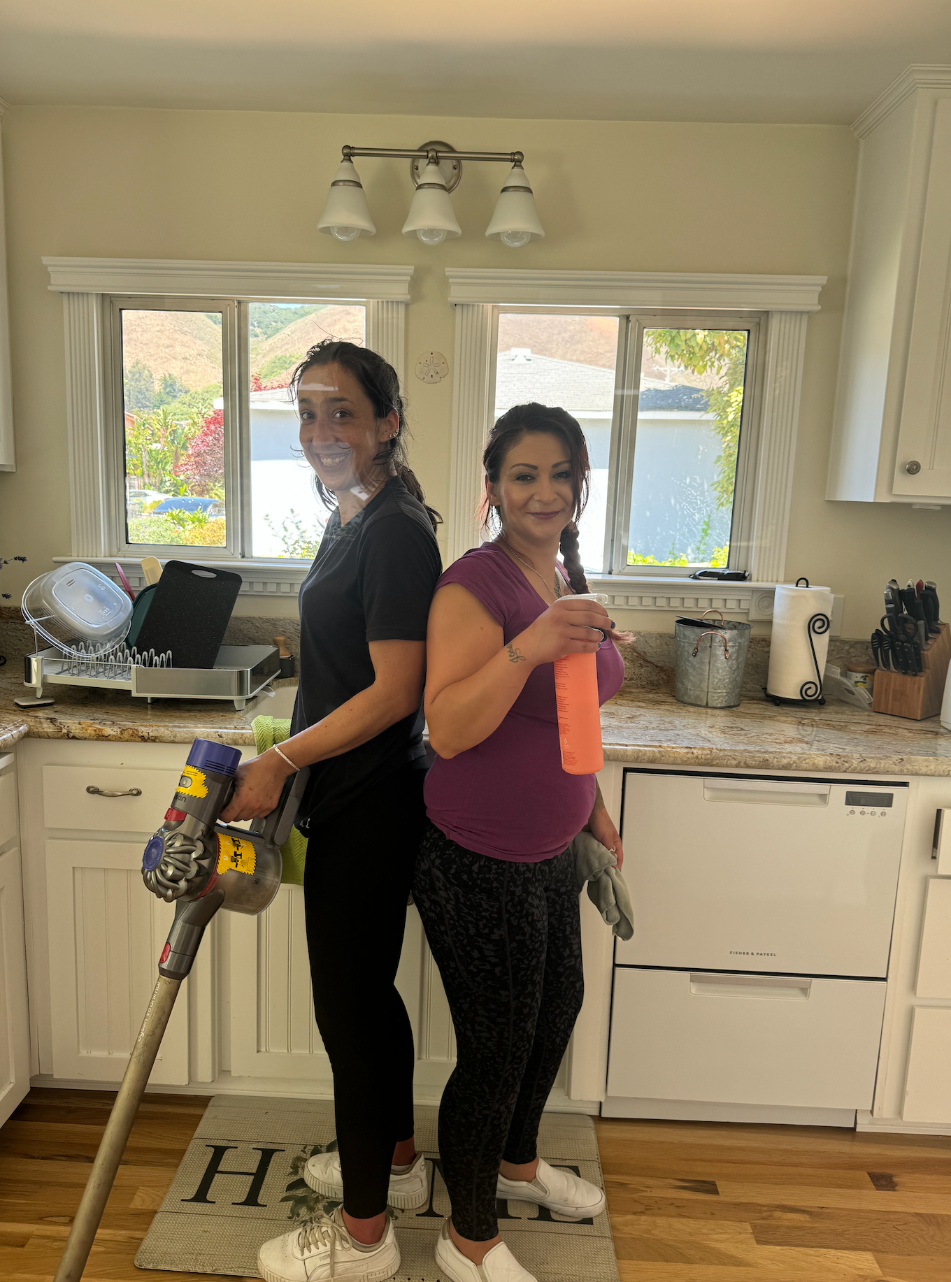Two women are standing next to each other in a kitchen holding cleaning supplies.