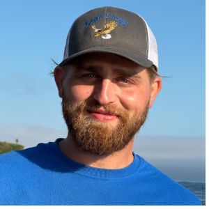 Man with a beard wearing a gray and white cap, smiling outside.