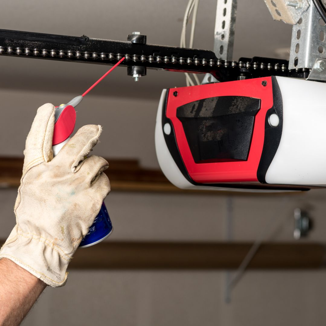 Gloved hand spraying lubricant on a garage door opener rail and moving trolley.
