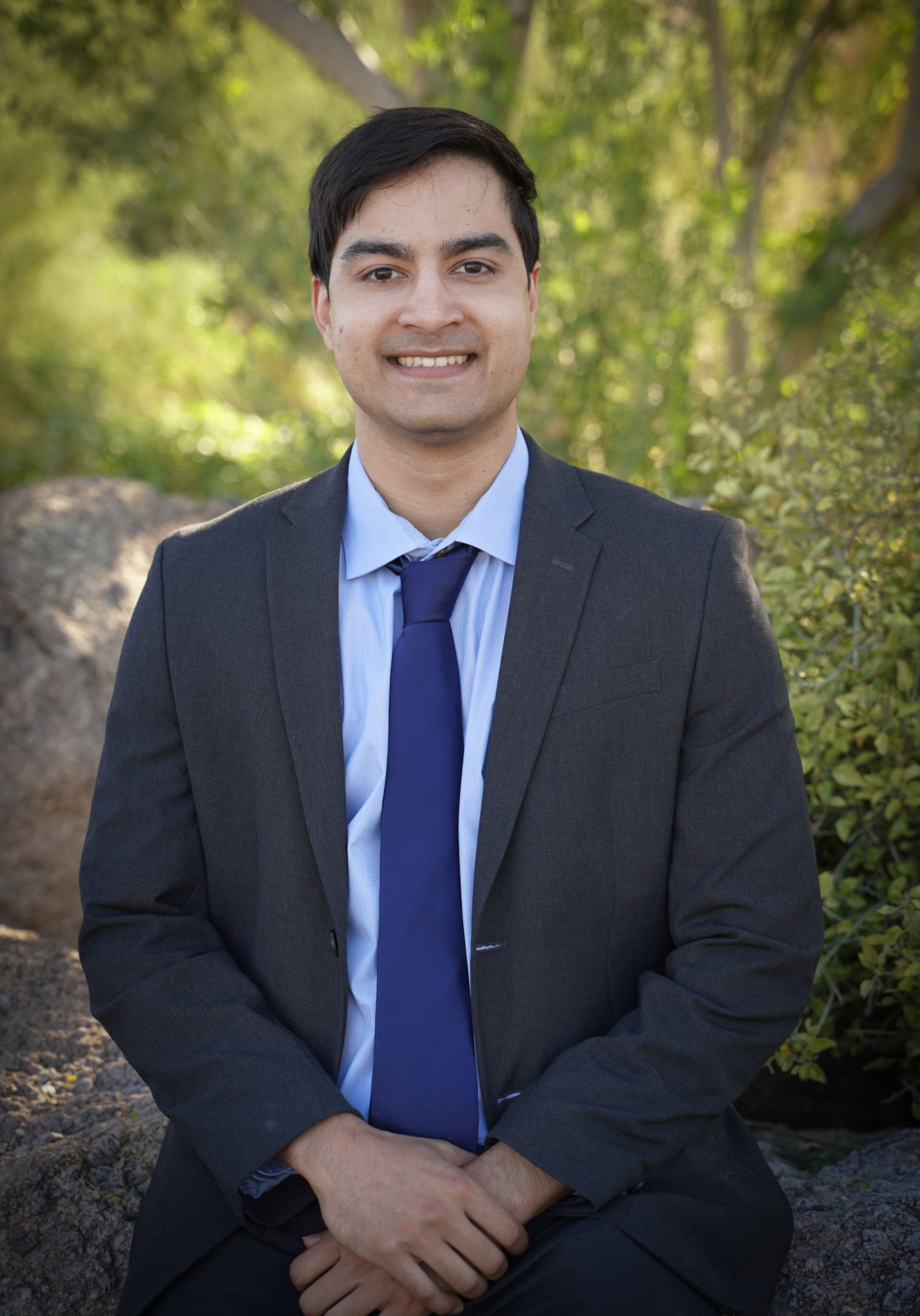 Man in suit smiles, seated outdoors. Dark suit, blue tie and shirt. Natural background.