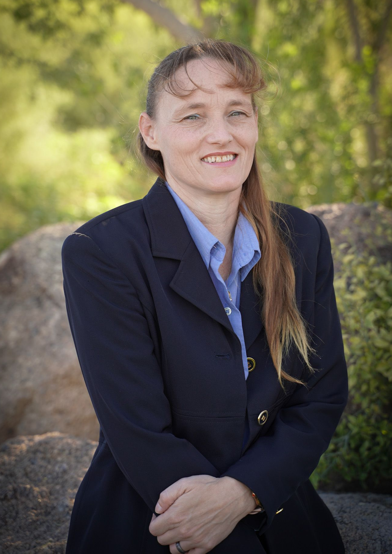Woman in blue blazer, button-down shirt smiles, arms crossed, outdoor setting with greenery.
