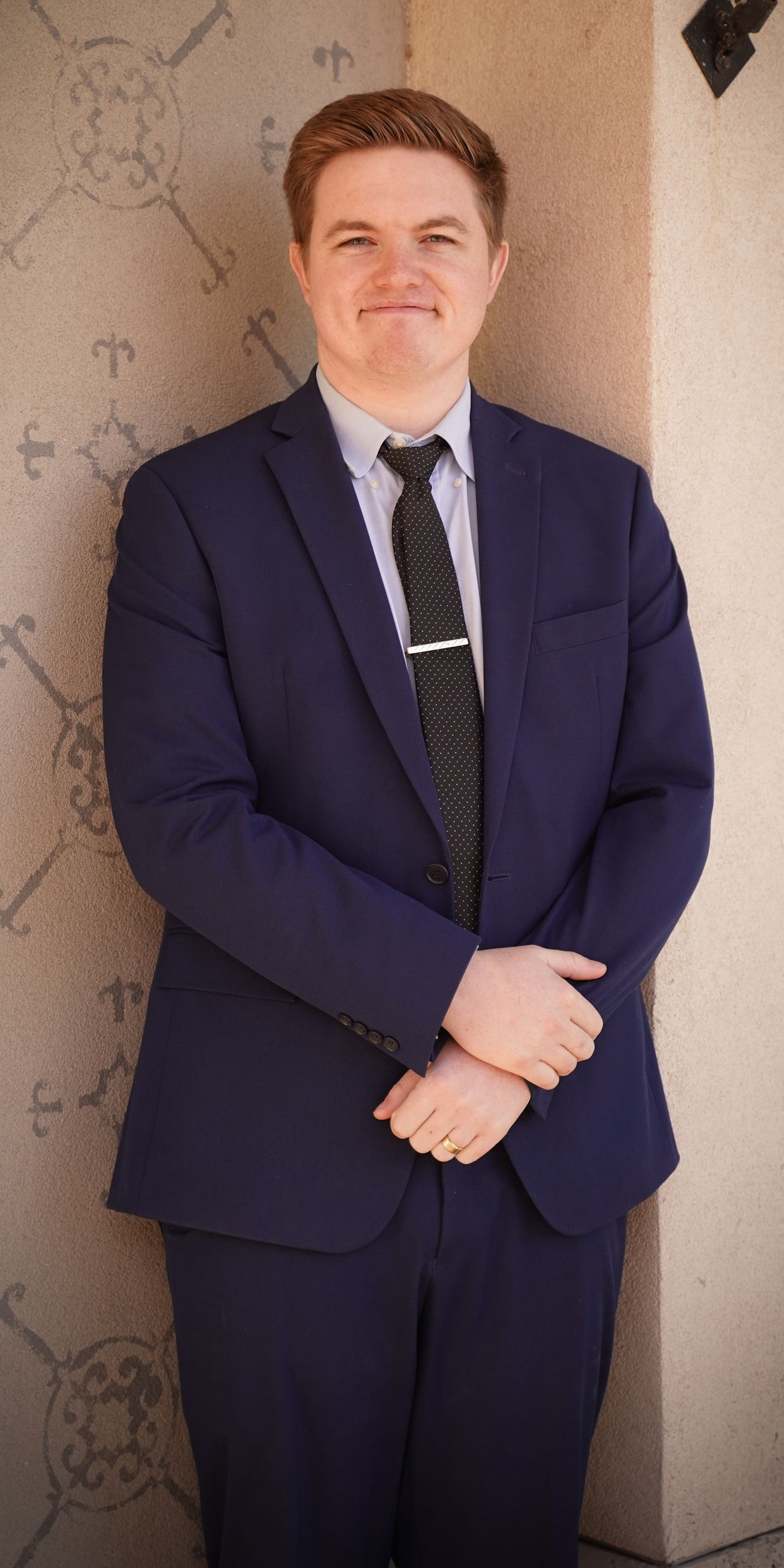 Man in a navy blue suit, standing near a beige wall with folded arms, smiling.