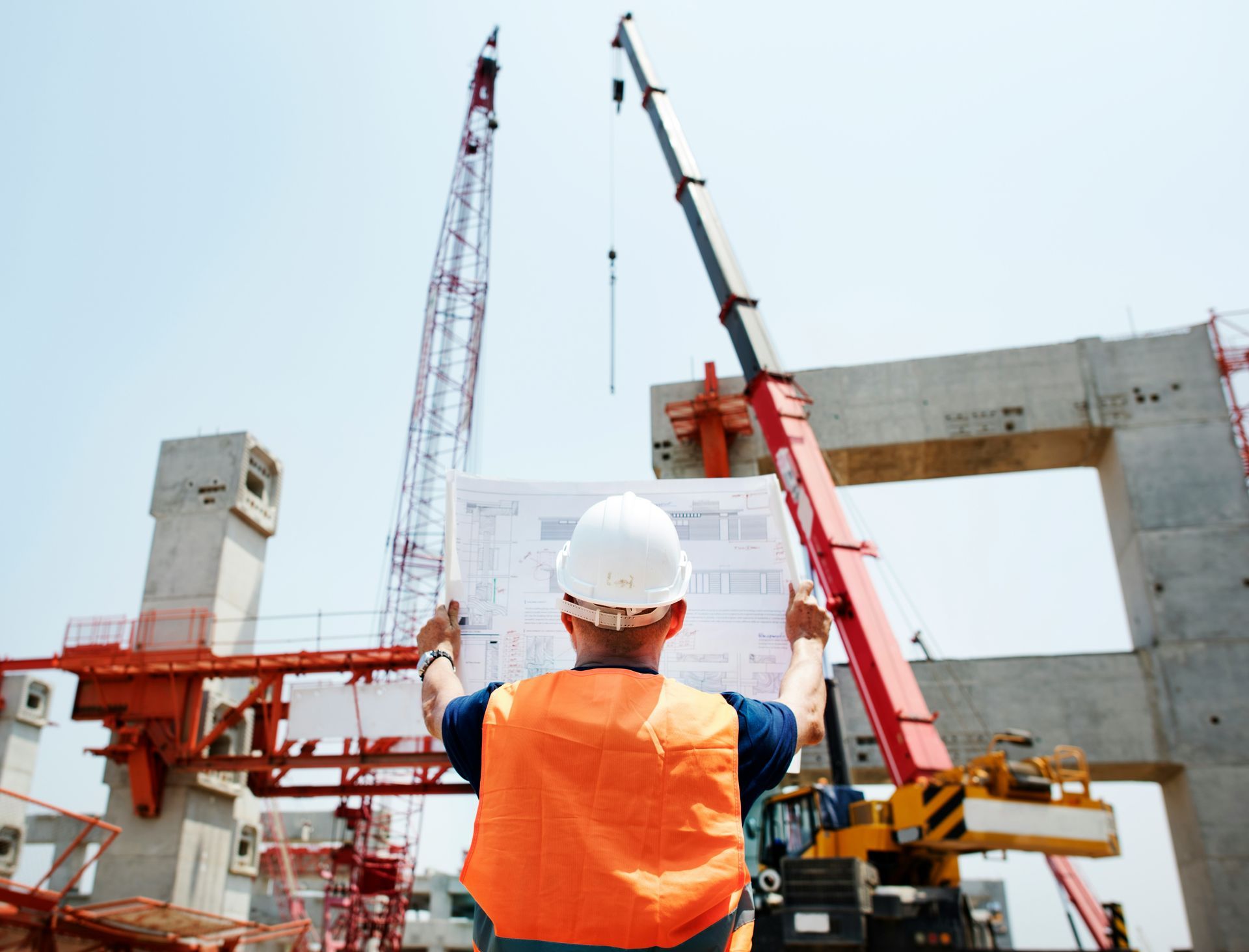 Construction worker in orange vest, looking at blueprints, cranes and concrete structure in background.