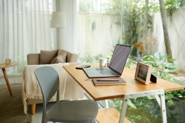 Home office with a laptop on a wooden desk. Light blue chair, sofa, and a window overlooking greenery.