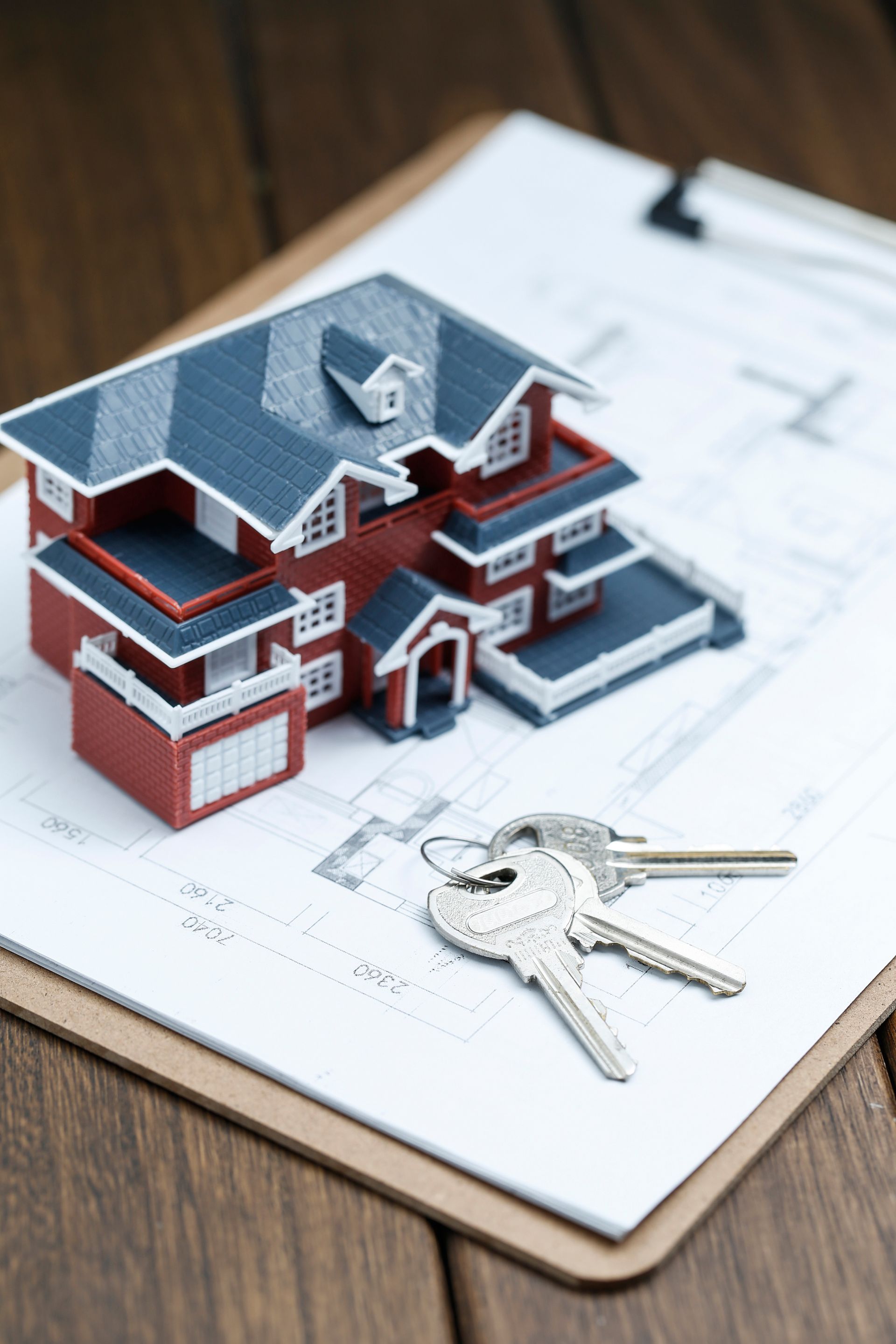 Model house, keys, and clipboard with blueprints on a wooden surface, representing a real estate transaction.