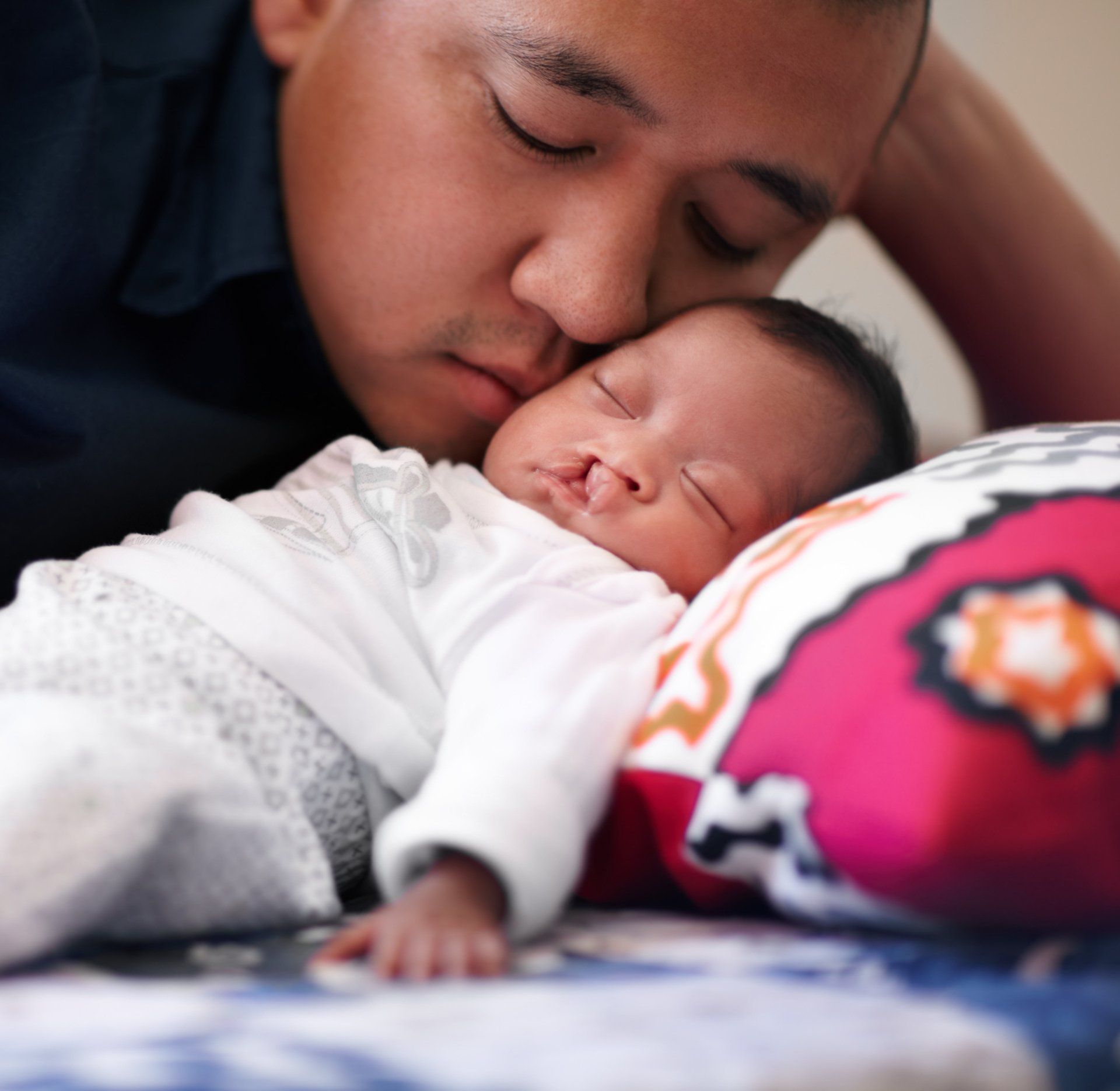 A parent with eyes closed gently presses their forehead against the cheek of a sleeping infant lying on a colorful pillow. Chet's Roof charity and community service seattle