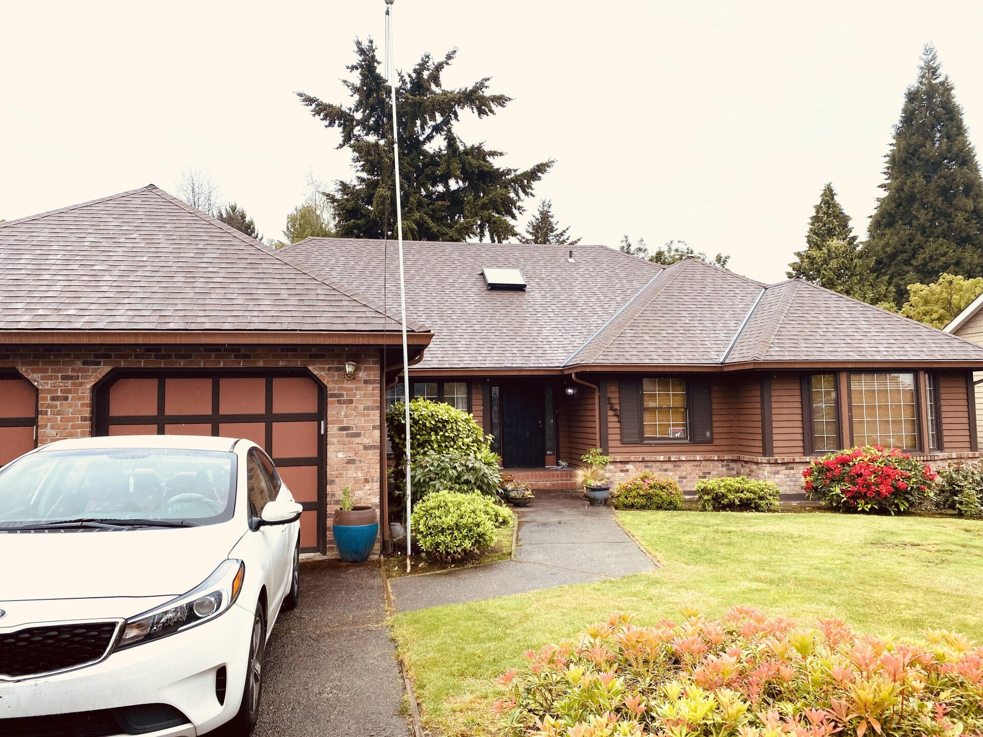 A single-story brown brick and wood house with a white car parked in the driveway and a green lawn in the front.