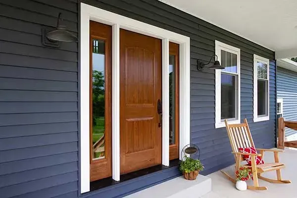 Blue house porch with wooden front door, white trim, and two wooden chairs on the right.