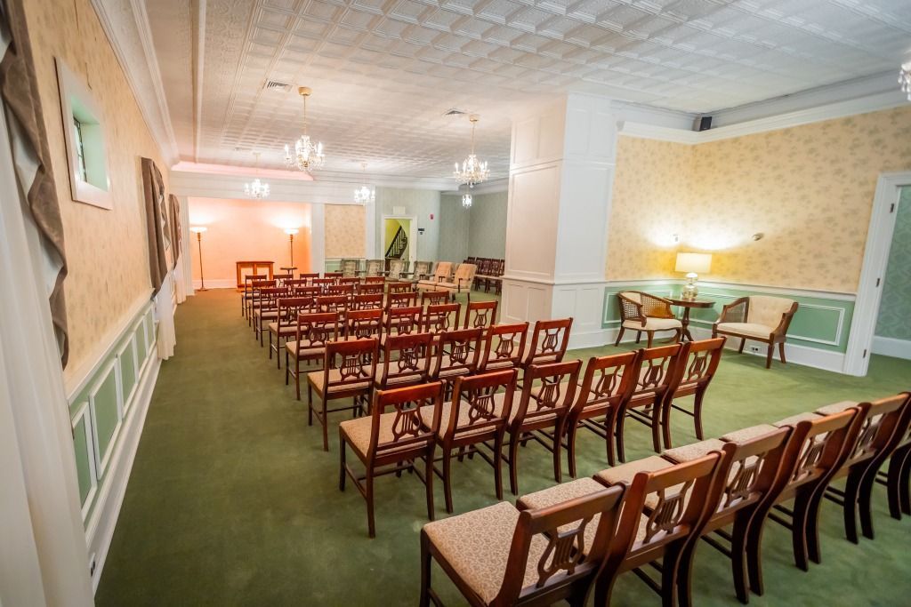 Rows of chairs face a small stage in a formal room, green carpet, decorative walls, chandeliers.