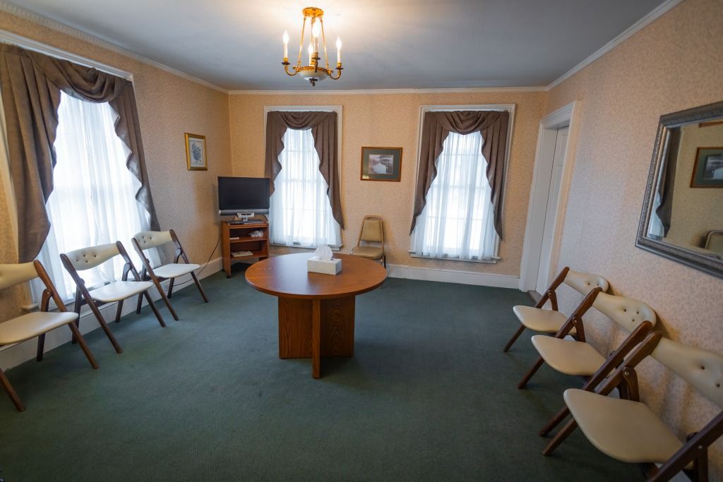 A room with chairs, a round table, windows with curtains, and a chandelier; possibly a waiting area.