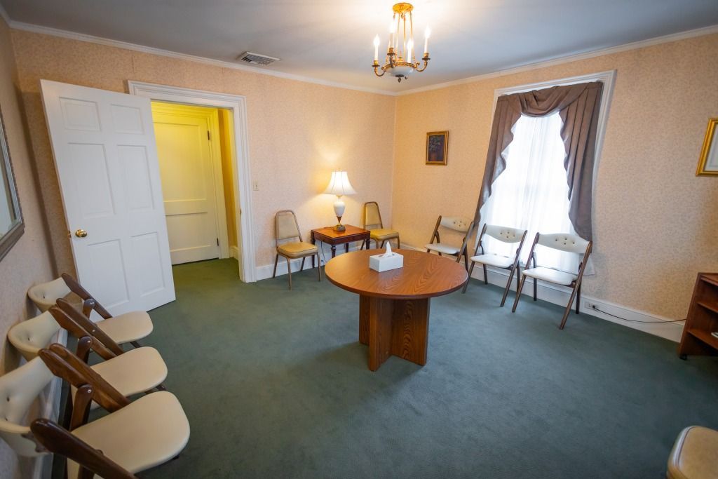 Waiting room with chairs, round table, and a window with curtains. Soft colors, carpeted floor.