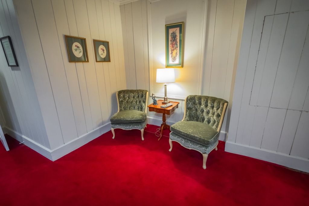 Two green velvet chairs facing each other beside a small table and lamp in a red-carpeted room.